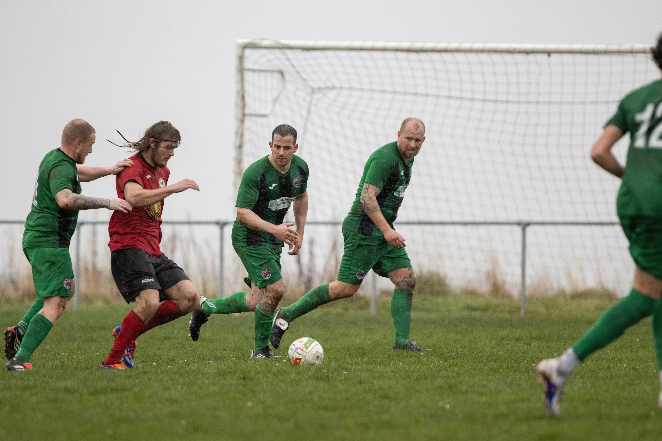 Soccer players in green and red jerseys competing for the ball on a grassy field near goalpost on overcast day.