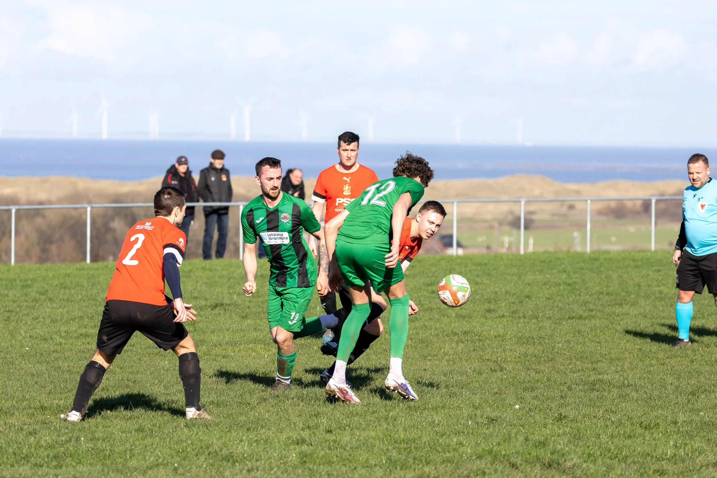 Soccer players in green and orange jerseys competing for the ball on a grassy field near the coast, with wind turbines visible in the background.