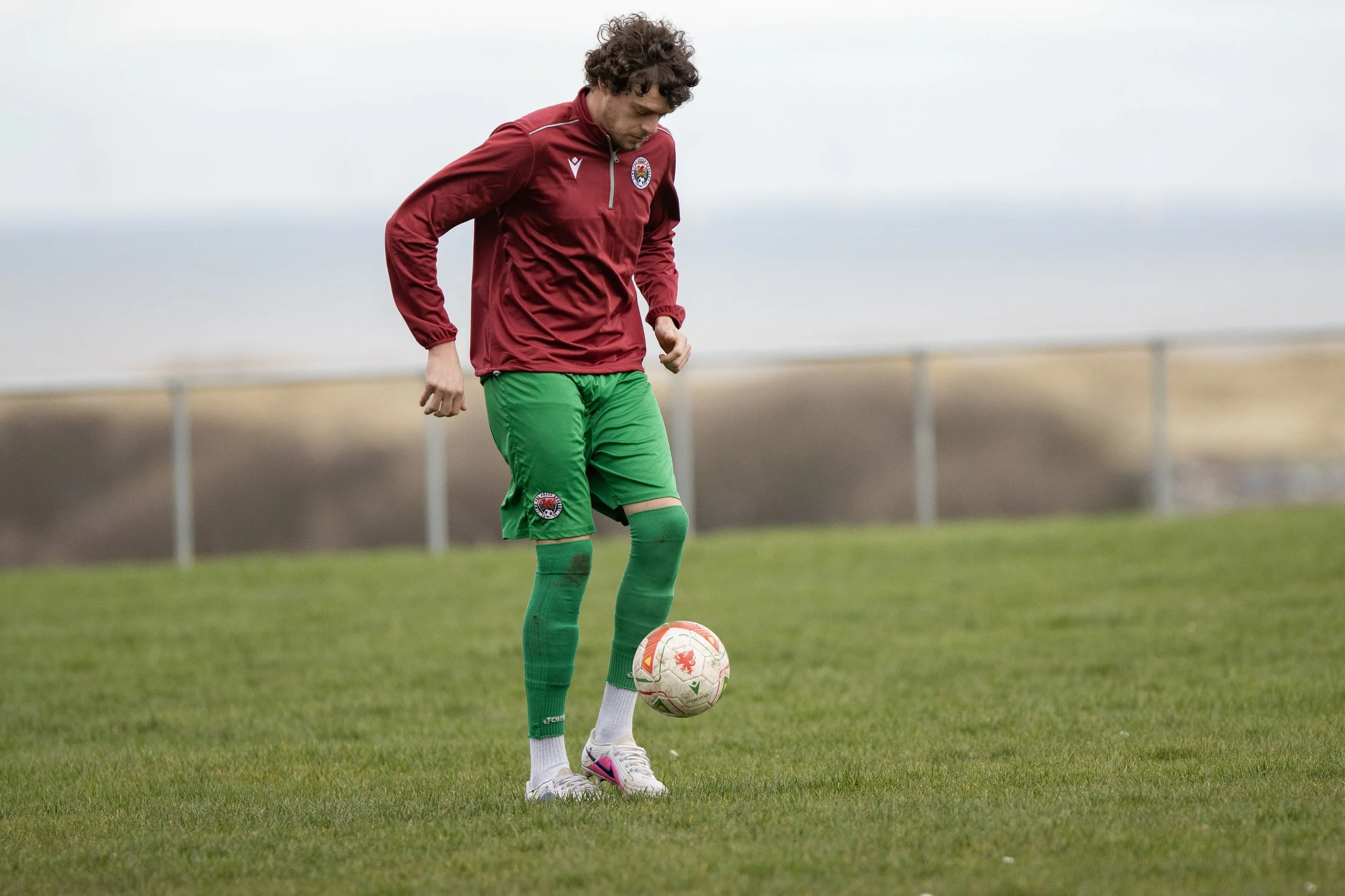 A soccer player wearing a maroon jacket, green shorts, and green socks, standing on a grassy field with a soccer ball at his feet.