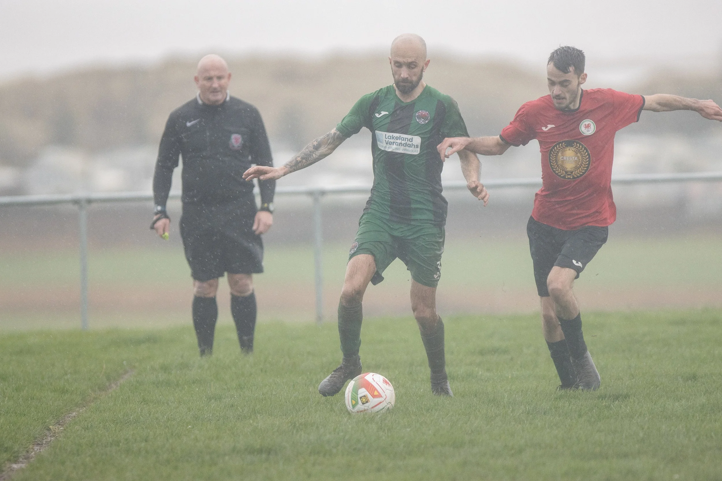 Two soccer players competing for ball on a rainy day, with a referee watching in the background.