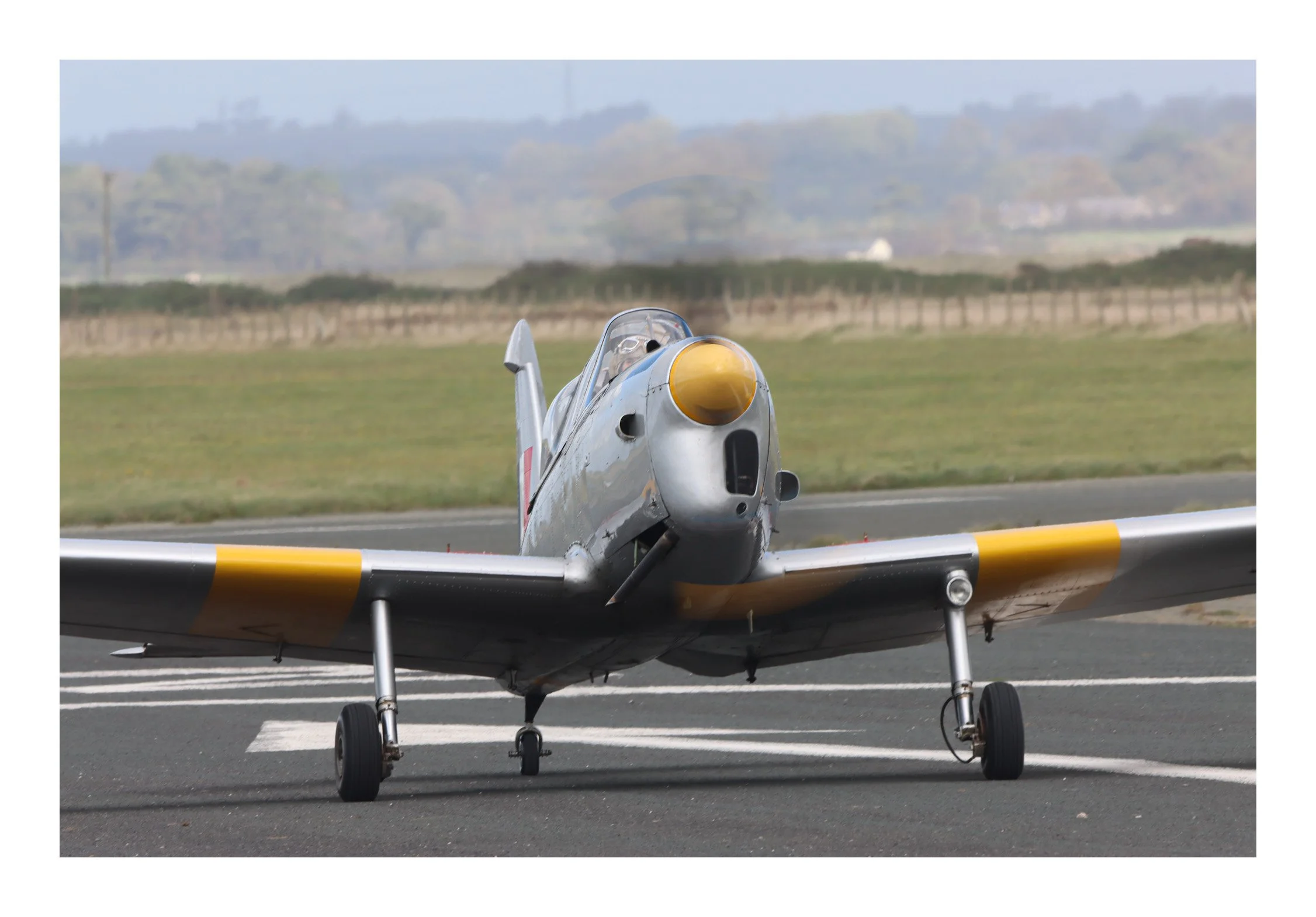 A vintage silver aircraft with yellow accents on the nose and wing tips, parked on a runway with grassy fields and distant trees in the background.