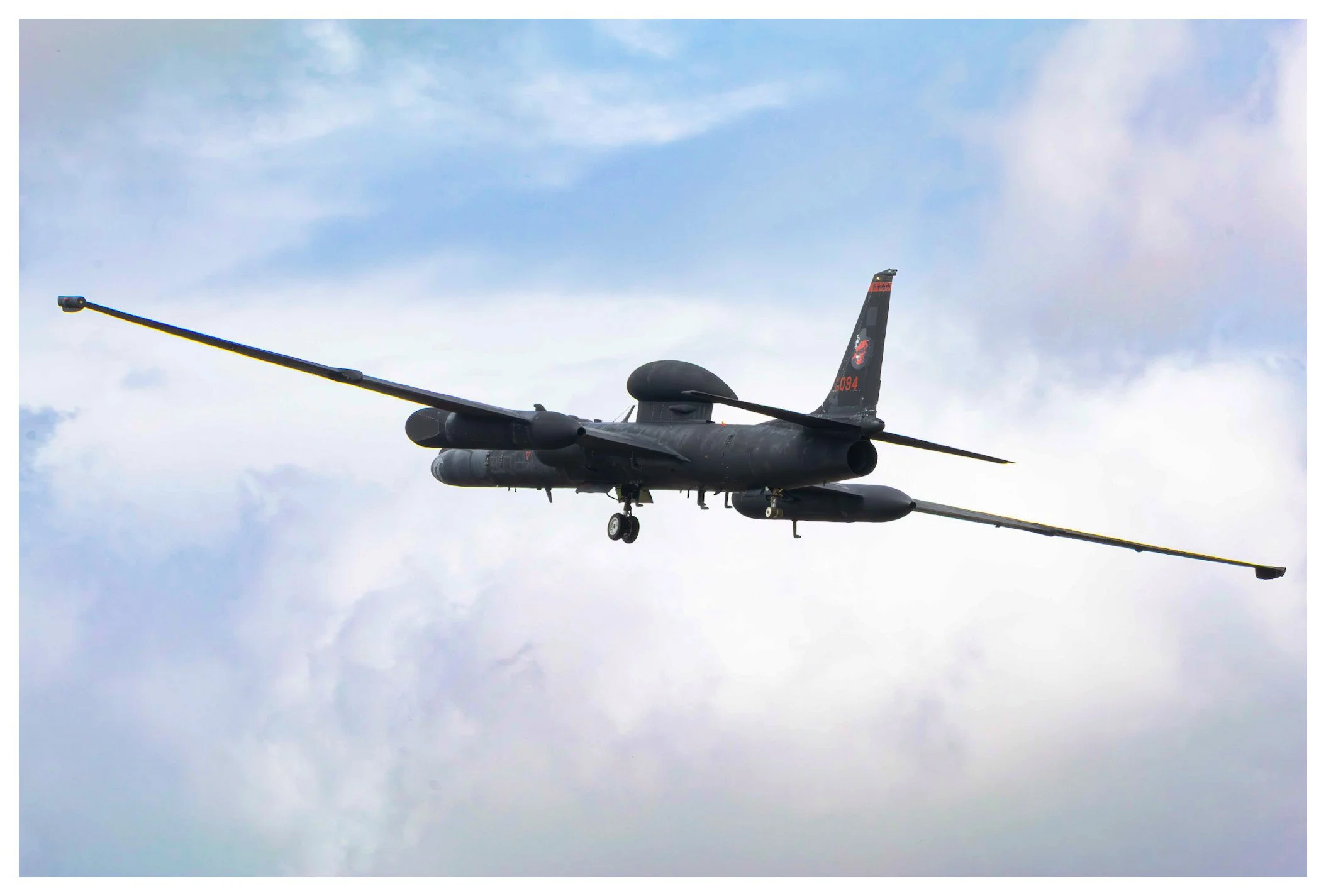 Black military drone flying in cloudy sky with extended wings and payload under the fuselage.