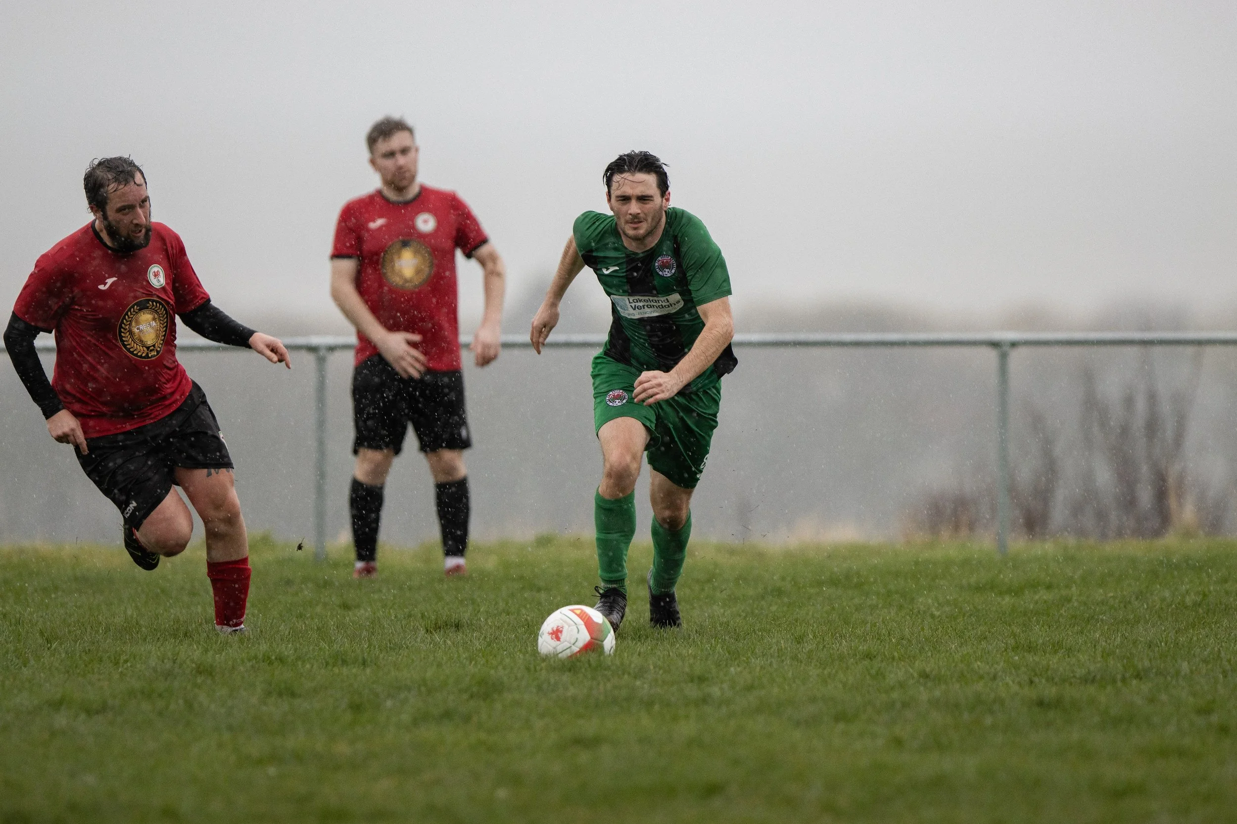 Three soccer players in rain, one in a green uniform dribbling a white ball with red and black details, two others in red and black uniforms chasing him on a grassy field.