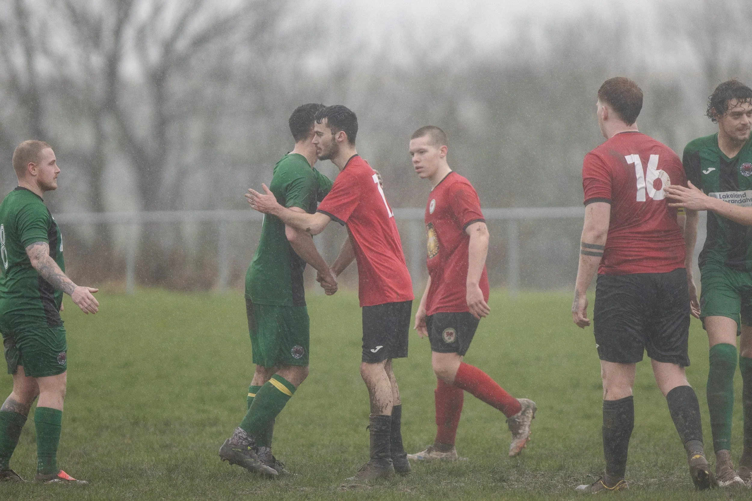 Soccer players in red and green uniforms shake hands on a rainy field after a match.