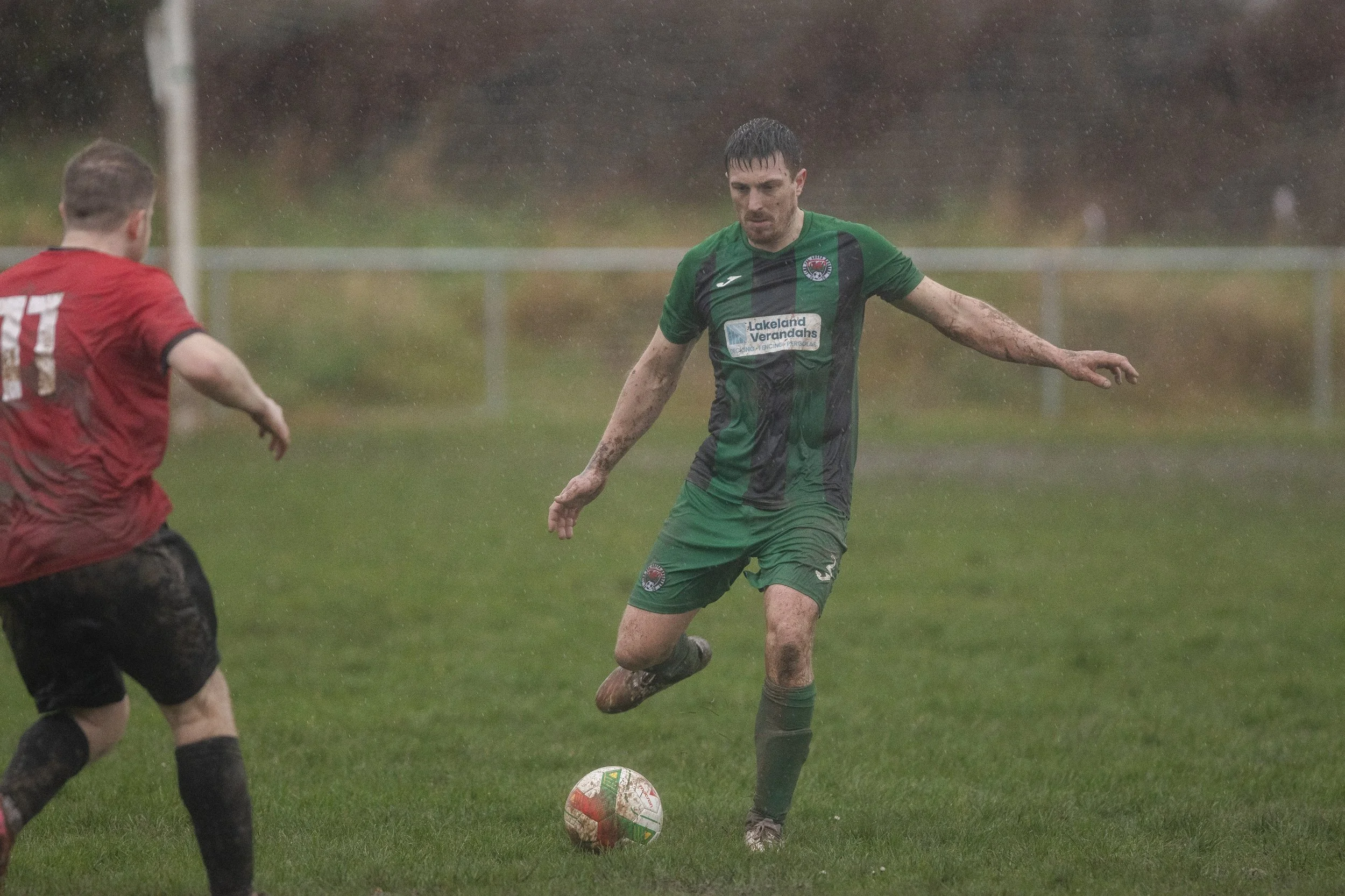 A muddy soccer game with two players, one in a green and black jersey and the other in a red jersey, playing on a wet grass field, rainy weather, with a blurred background of trees and a fence.