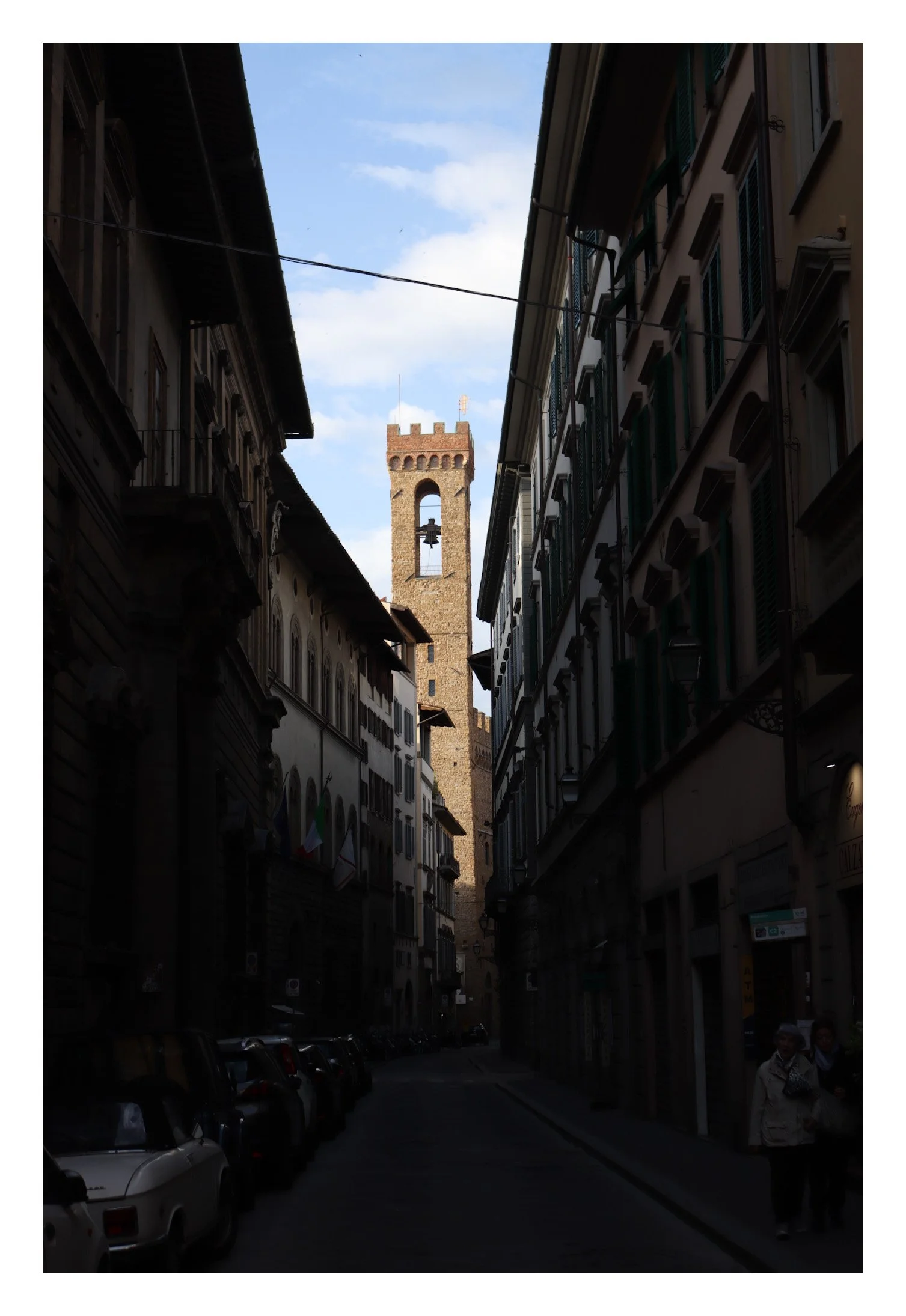 A narrow street in an old European city with tall, historic buildings on both sides, leading to a brick clock tower with a bell under a blue sky with scattered clouds.