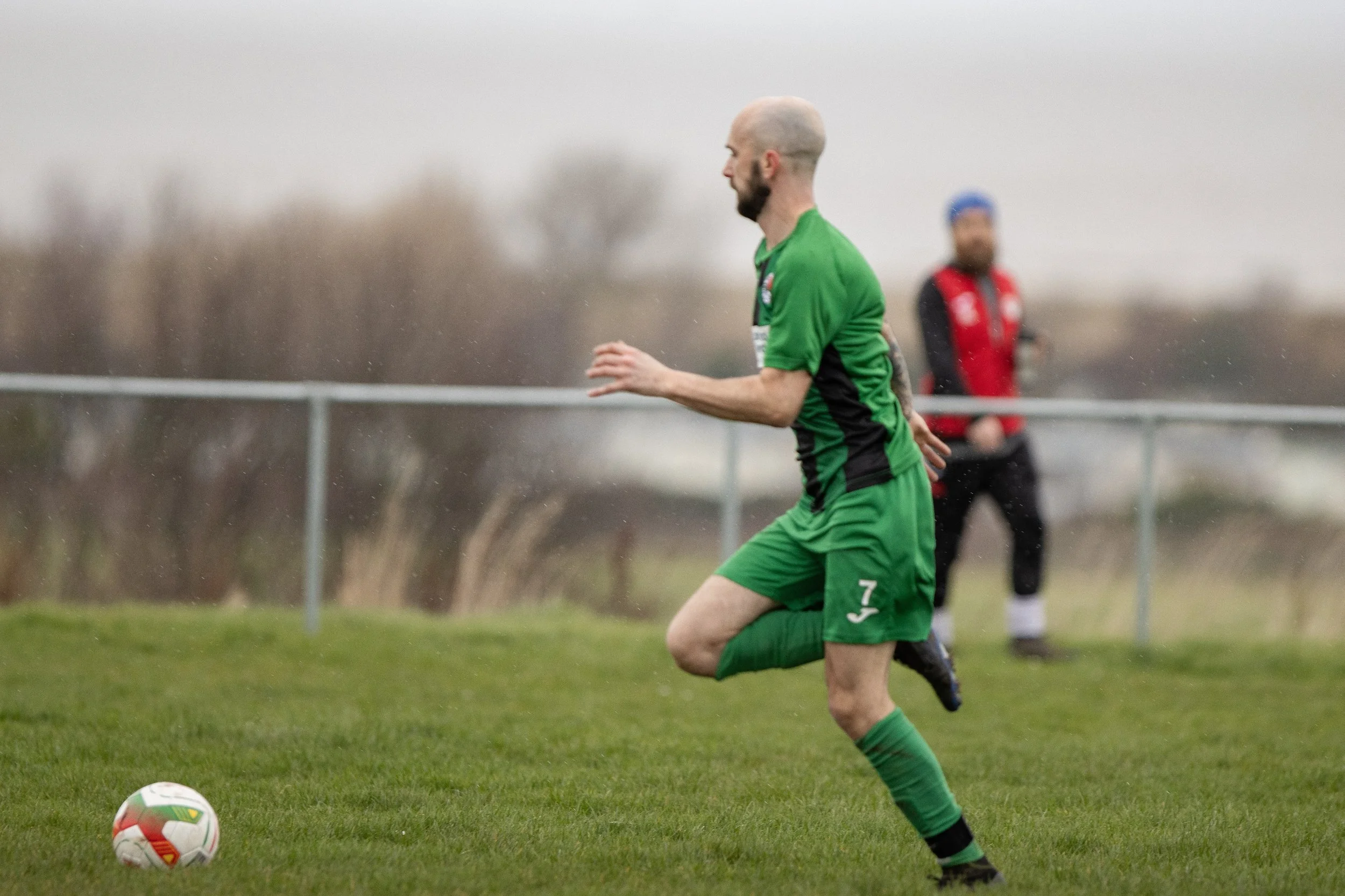 A man in a green soccer uniform running on a grassy field during a cloudy day, with a soccer ball in front of him.