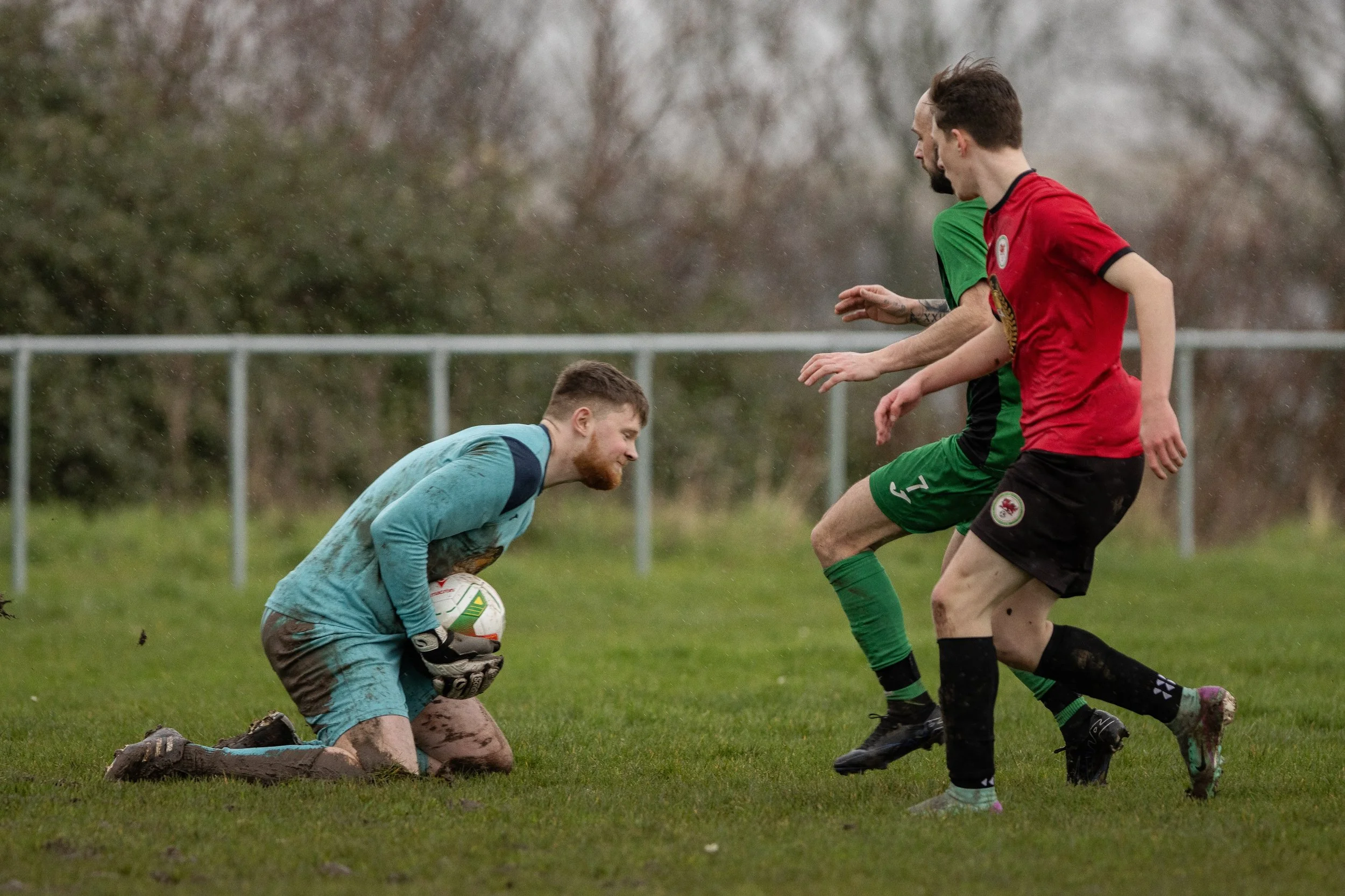 A soccer goalkeeper in a teal kit kneeling on the grass, holding a soccer ball, facing two players from the opposing team in red and green jerseys during a match in rainy weather.