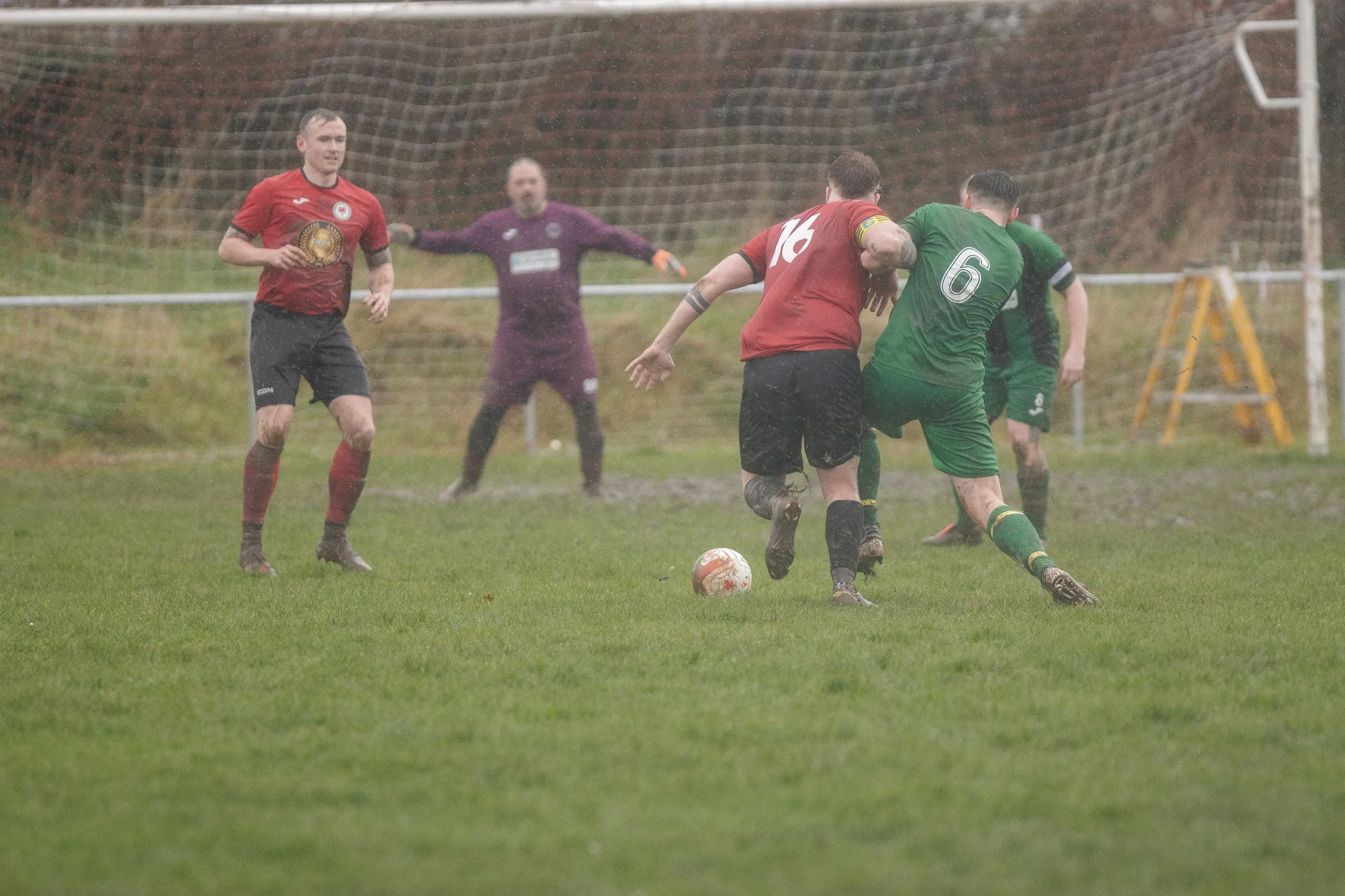 Soccer players in a match during rain, with one player in a red jersey and another in a green jersey fighting for the ball, while other players and a goalkeeper watch.