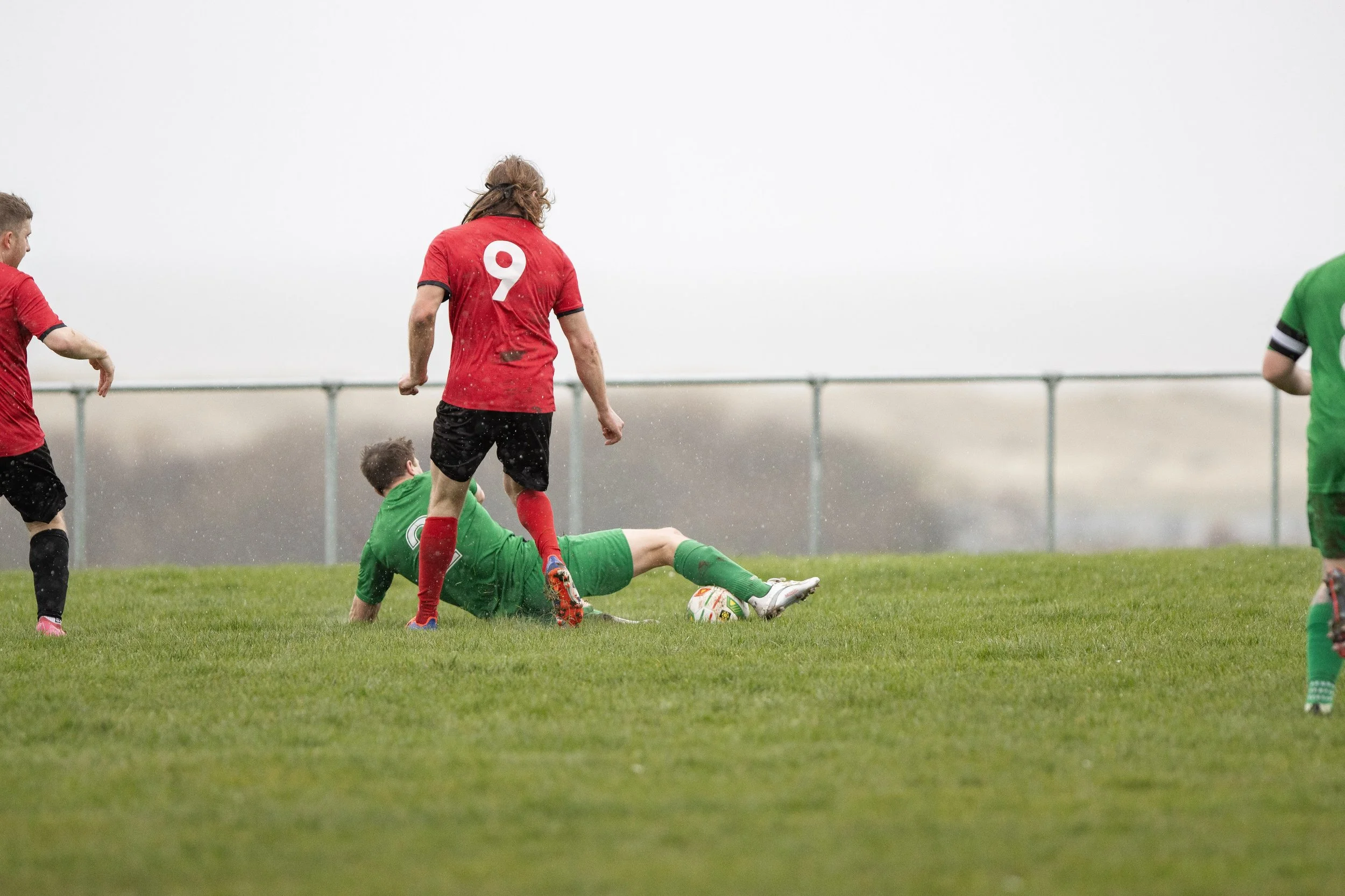 Soccer players on a wet field during a game, with one player in green lying on the ground and others in red nearby.