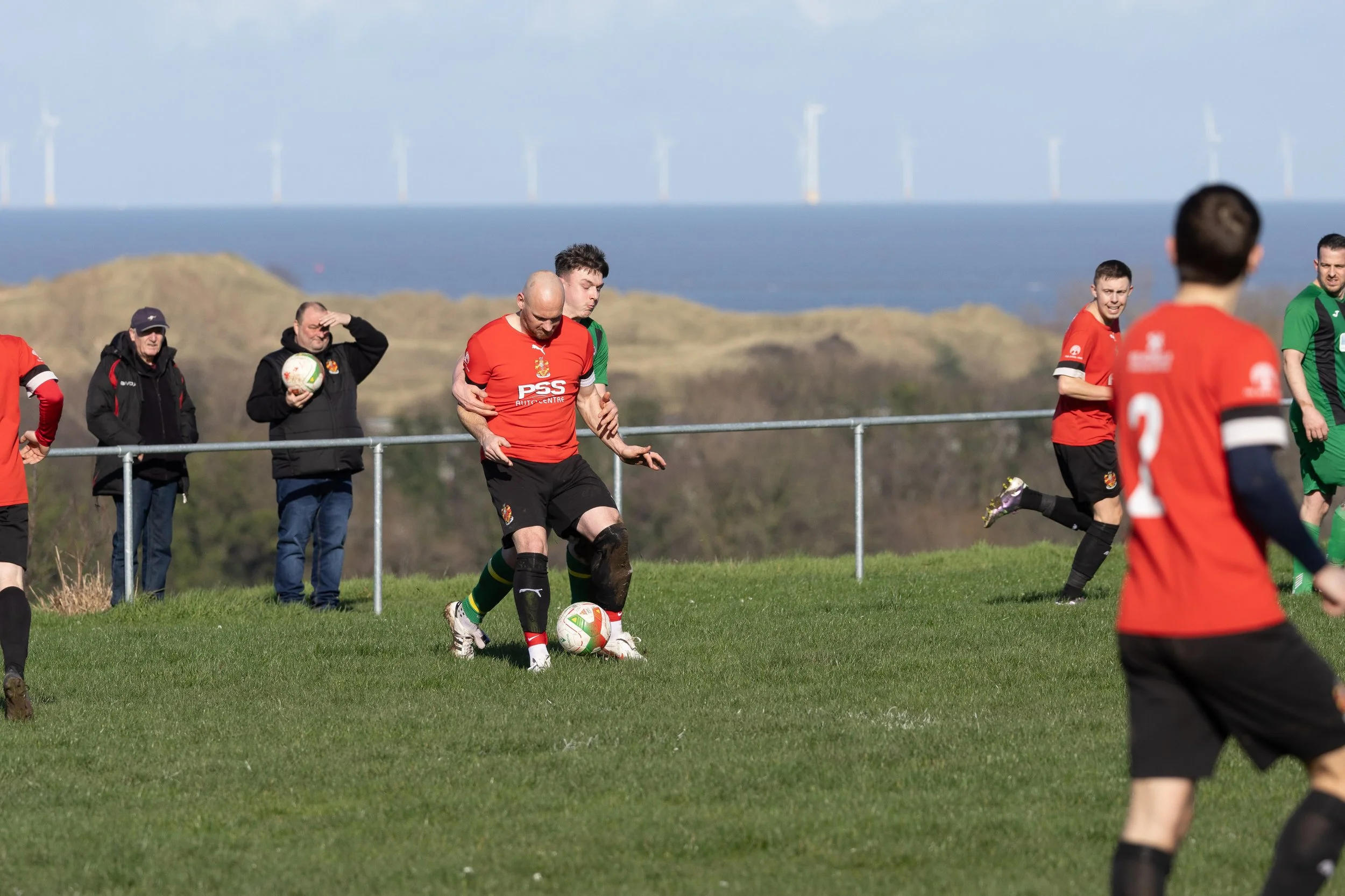 Soccer players on a grassy field, some wearing red jerseys and black shorts, playing during a match at outdoors with a coastline and wind turbines in the background.