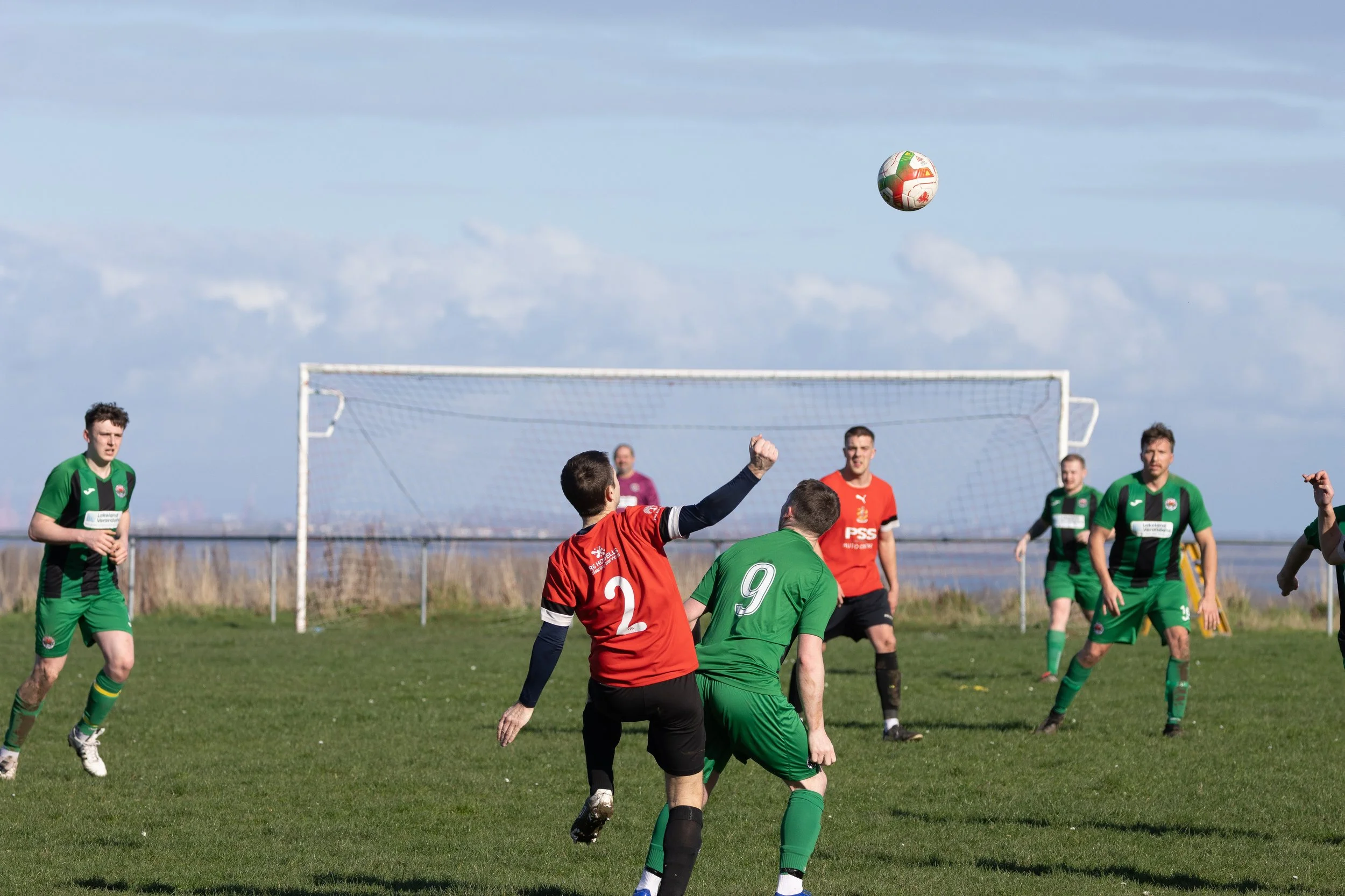 Soccer players in green and red jerseys on a grassy field during a match, with a goalpost and soccer ball in the air.