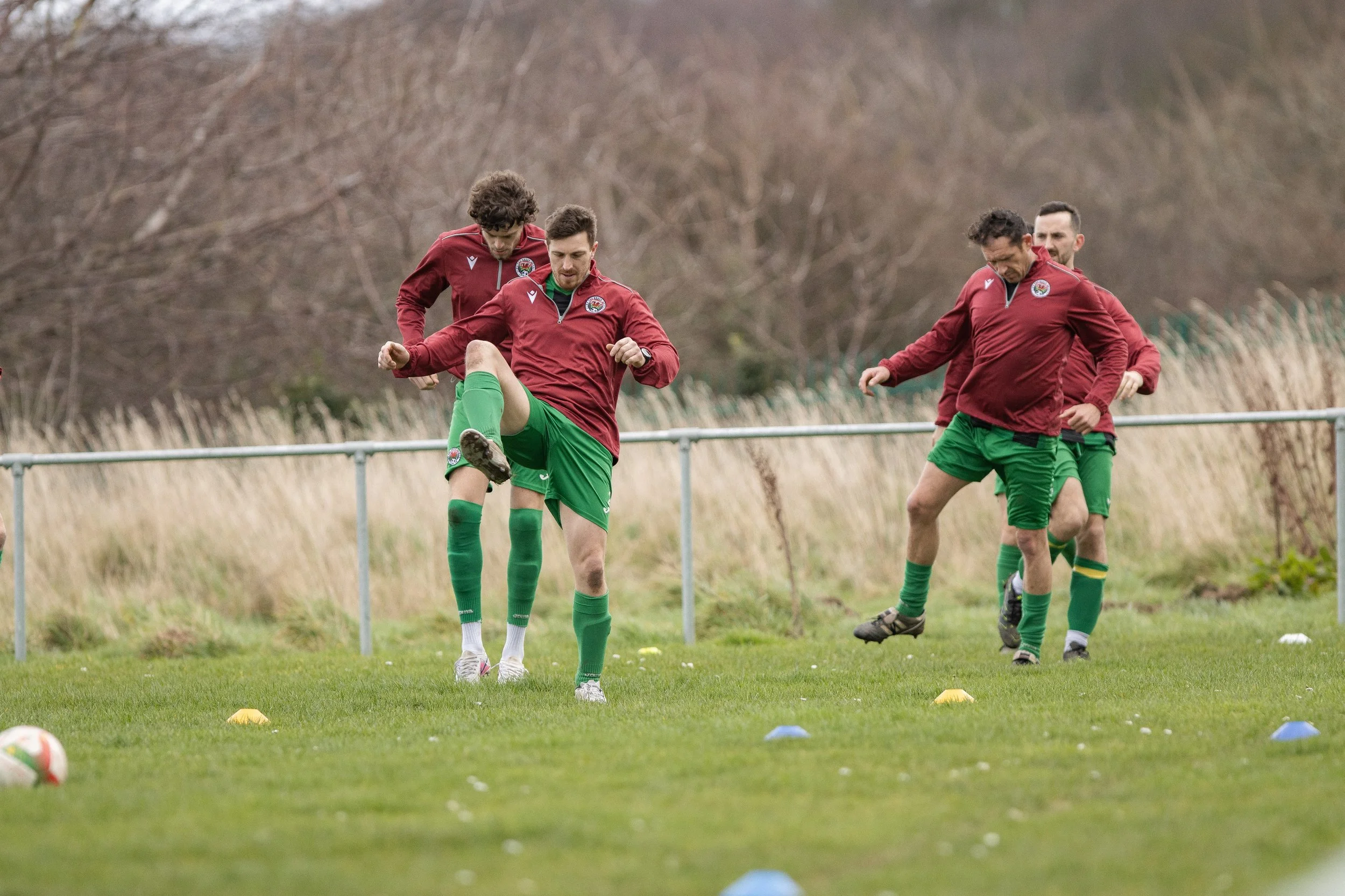Group of male soccer players in red jackets and green shorts practicing on a grassy field, warming up with drills.