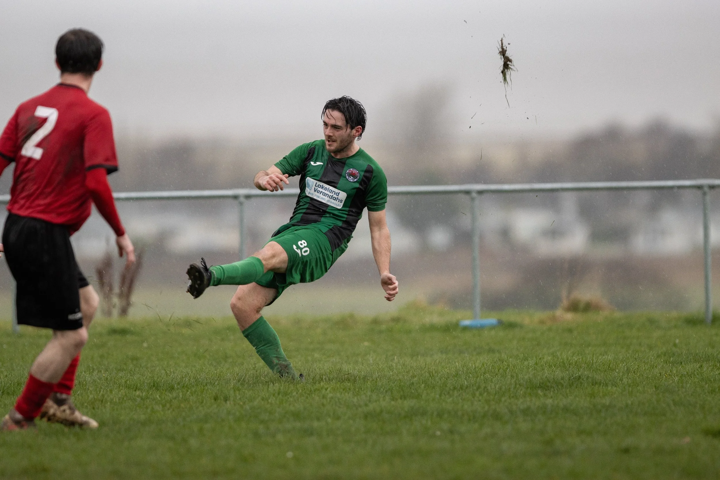 A soccer player in a green uniform is kicking a soccer ball while another player in a red uniform watches on a rainy day.