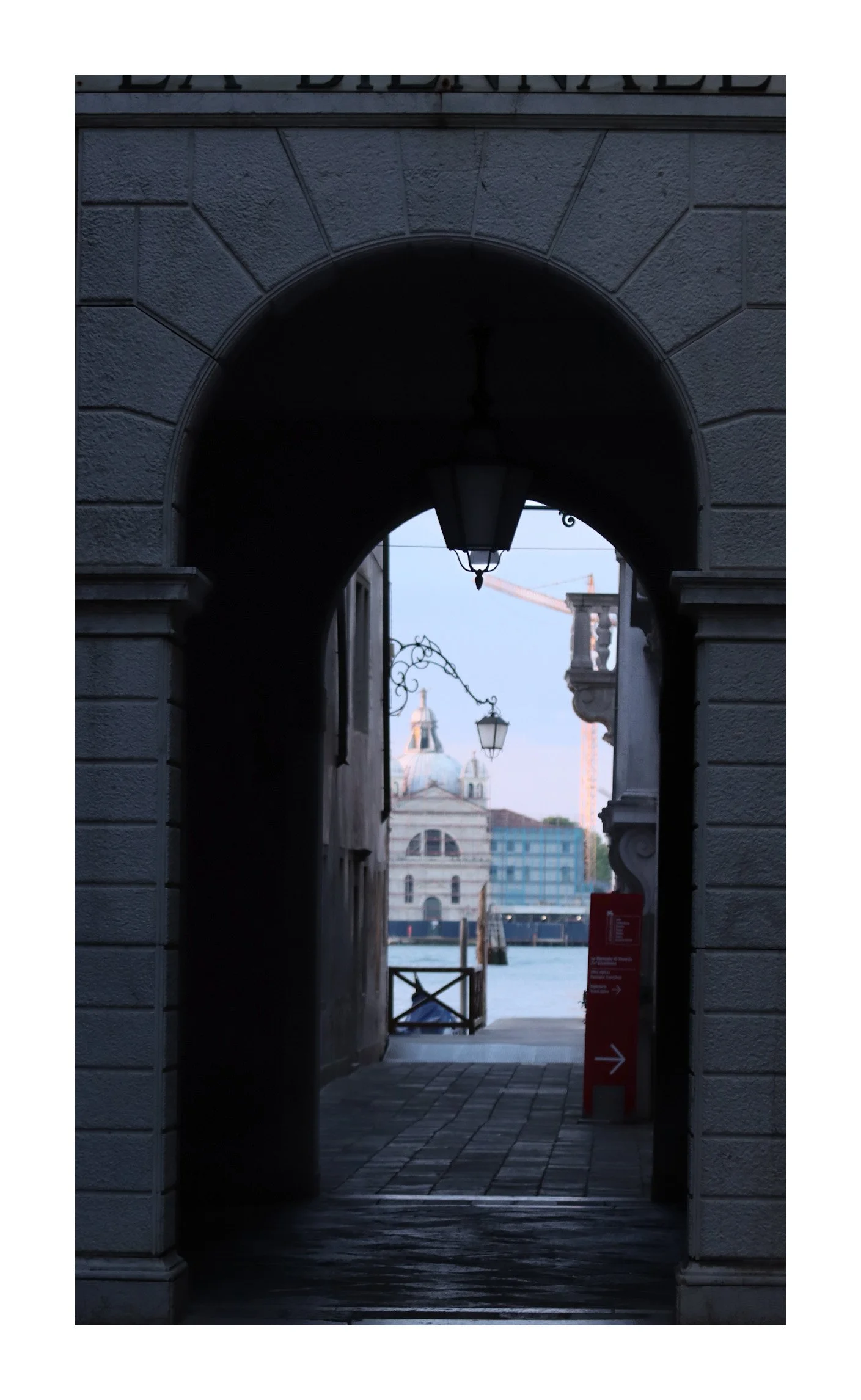 View through arched gate revealing church with dome and spire across water, with construction cranes and modern buildings in background.