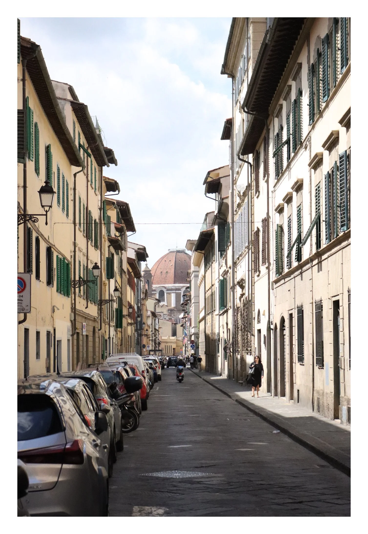 A narrow street lined with parked cars and multi-story buildings with green shutters, leading to a domed church in the background, in an Italian city.