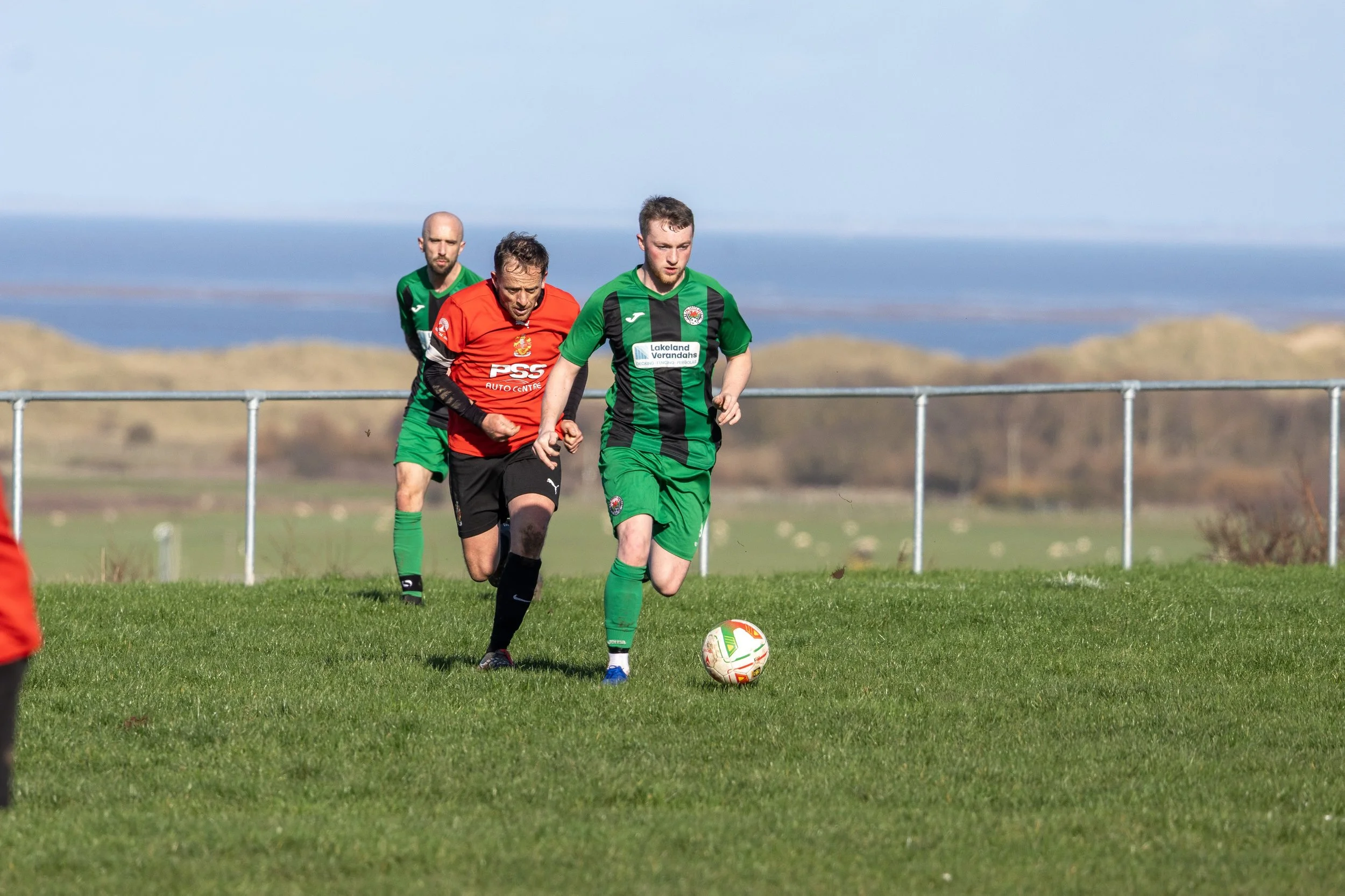 Soccer players on the field, one in green and black uniform dribbling the ball, others in red and black uniforms chasing him, on a grassy field with a fence and hills in the background.