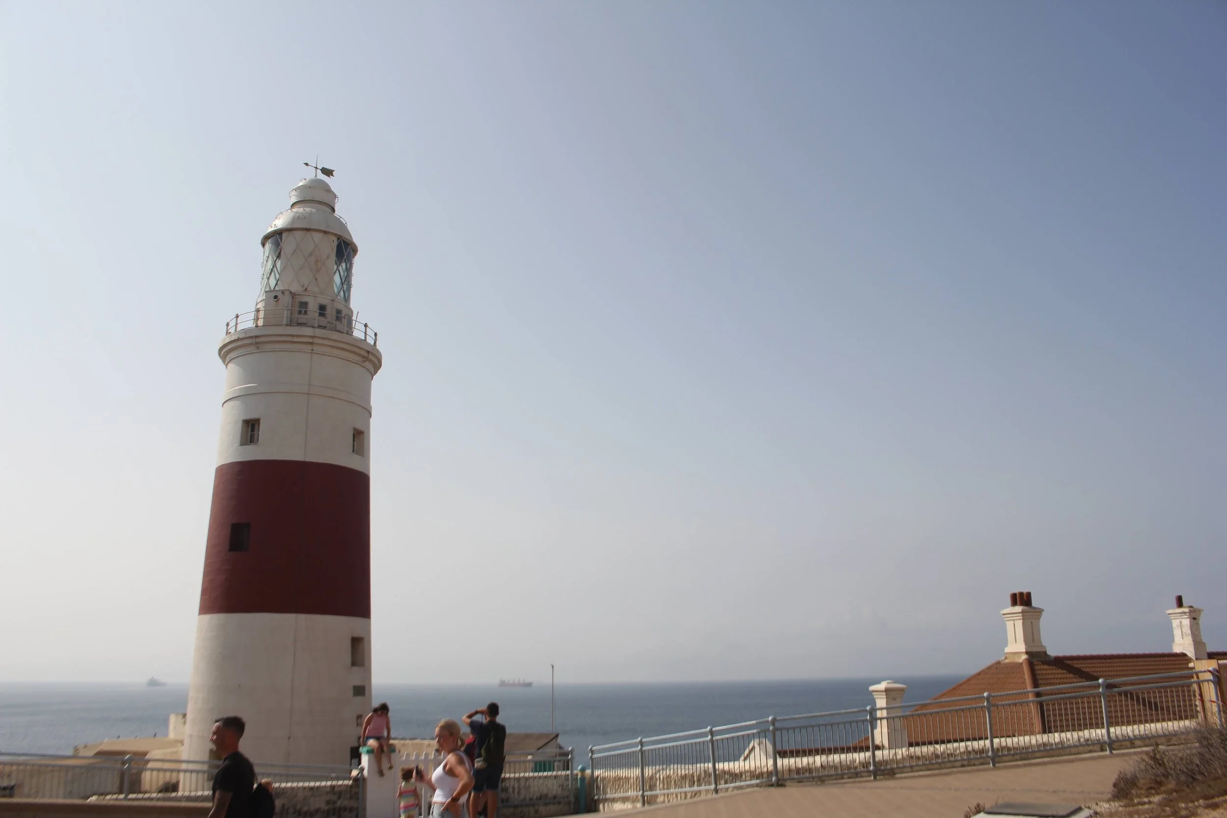 A white and red striped lighthouse by the sea with people walking nearby and ships in the distance.