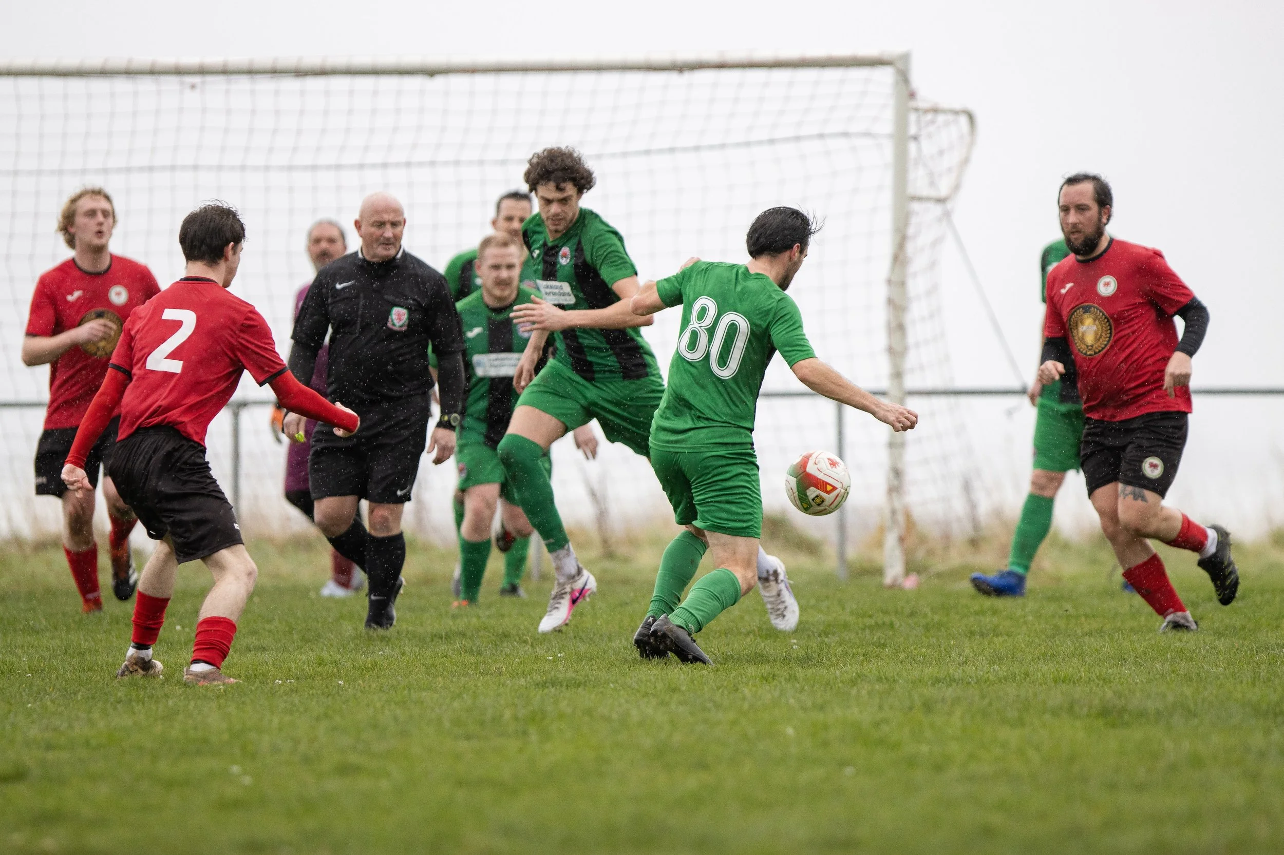 Soccer game with players in green and red uniforms chasing the ball near the goal, with a referee nearby on a grassy field on a cloudy day.