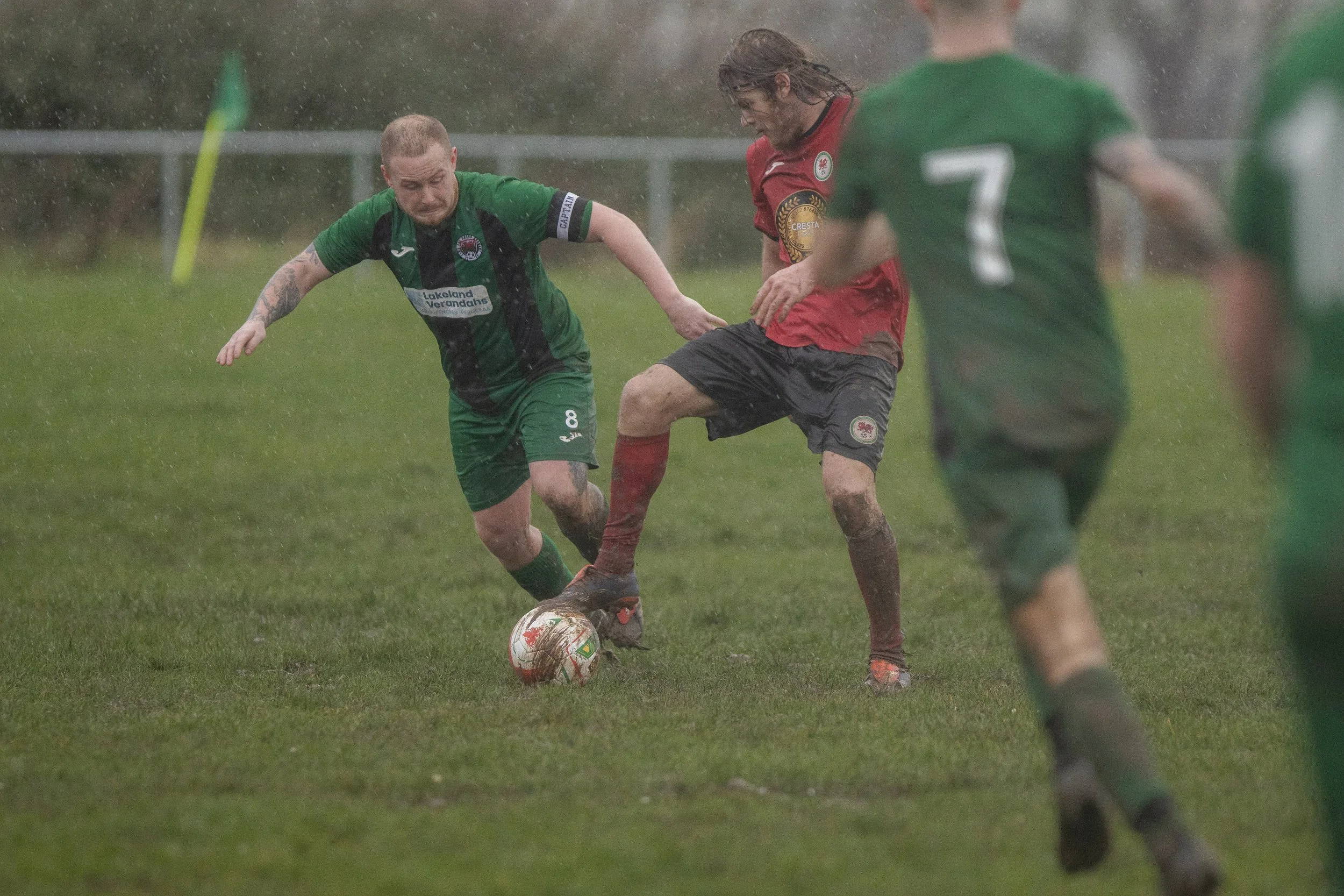 Soccer players in a rainy match competing for the ball on a muddy field, wearing green and red jerseys.