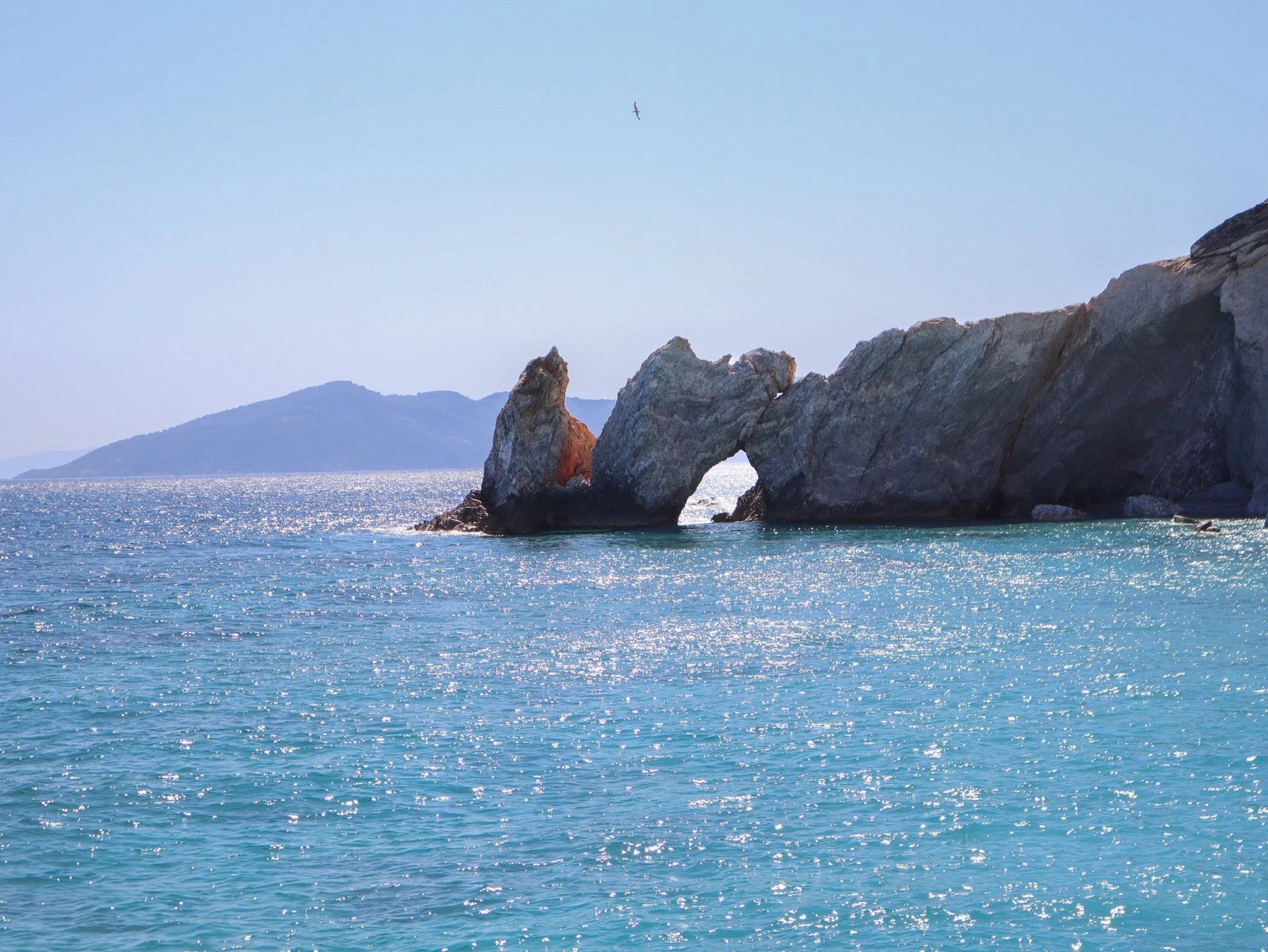 A coastal scene with a large natural rock formation featuring a hole, turquoise water, a distant island, and a bird flying in the sky.