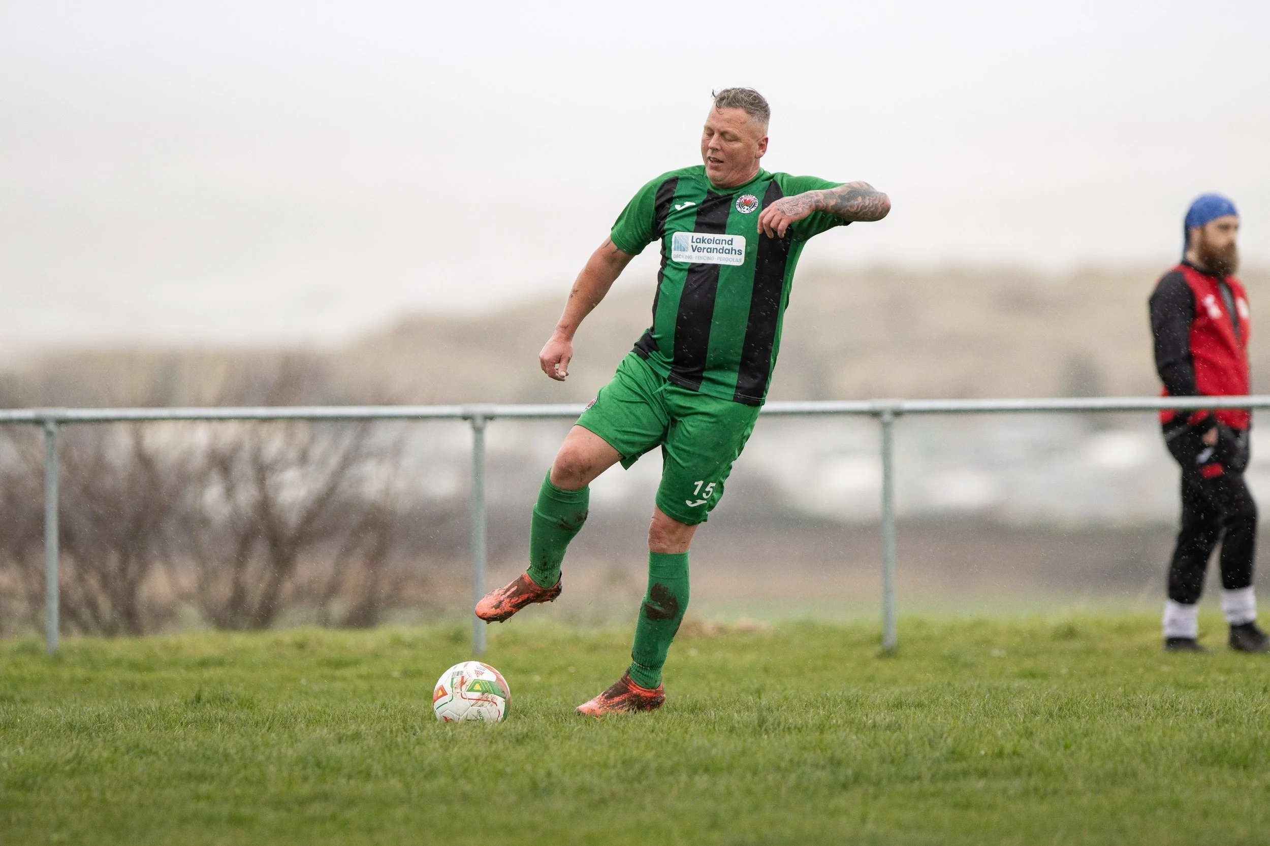 A soccer player in a green and black uniform, kicking a soccer ball on a grassy field during a match on a cloudy day.