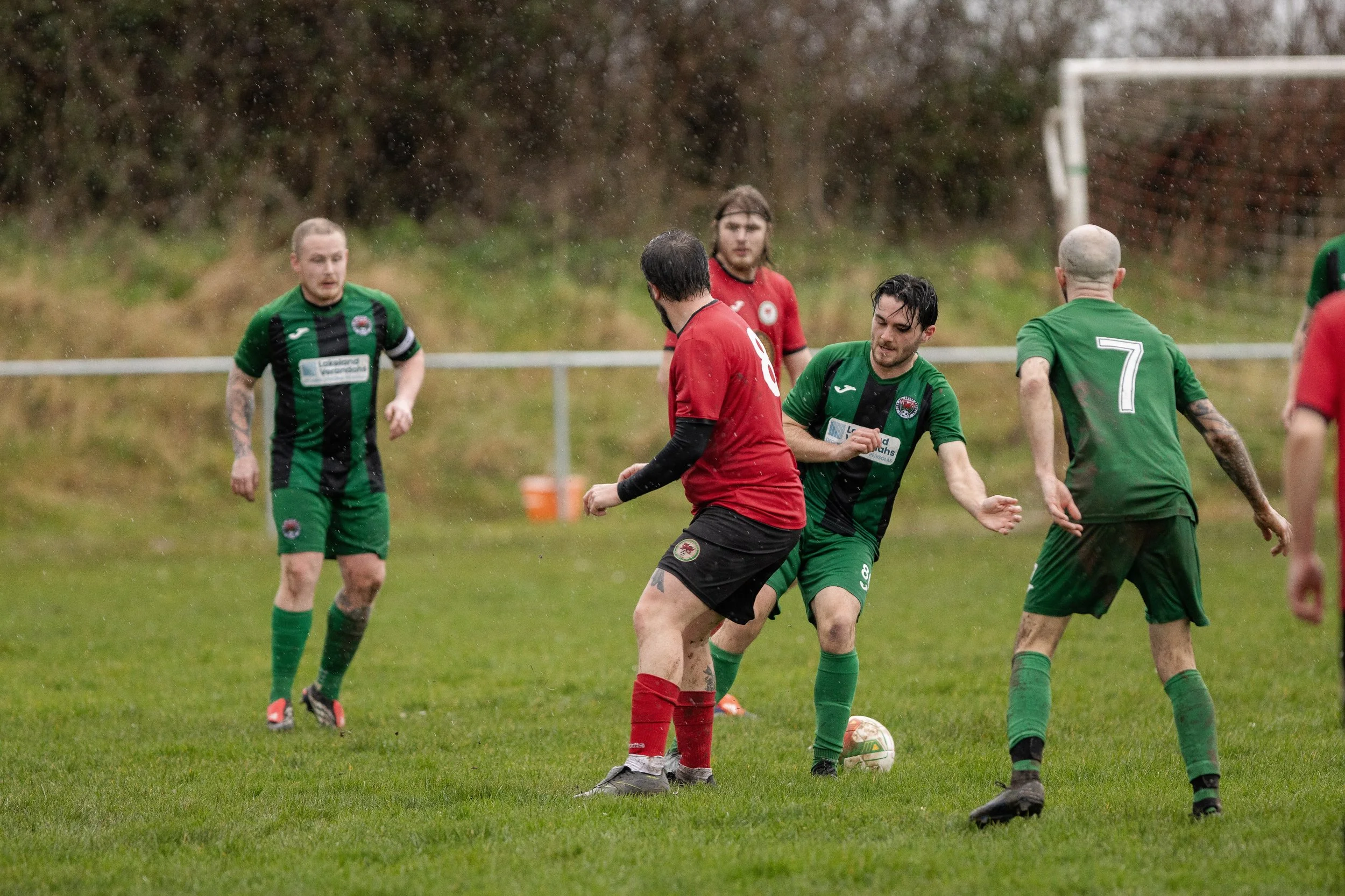Soccer players on a field during a match, with rain falling. Five players are visible, wearing green and red uniforms, actively engaged near the soccer ball.