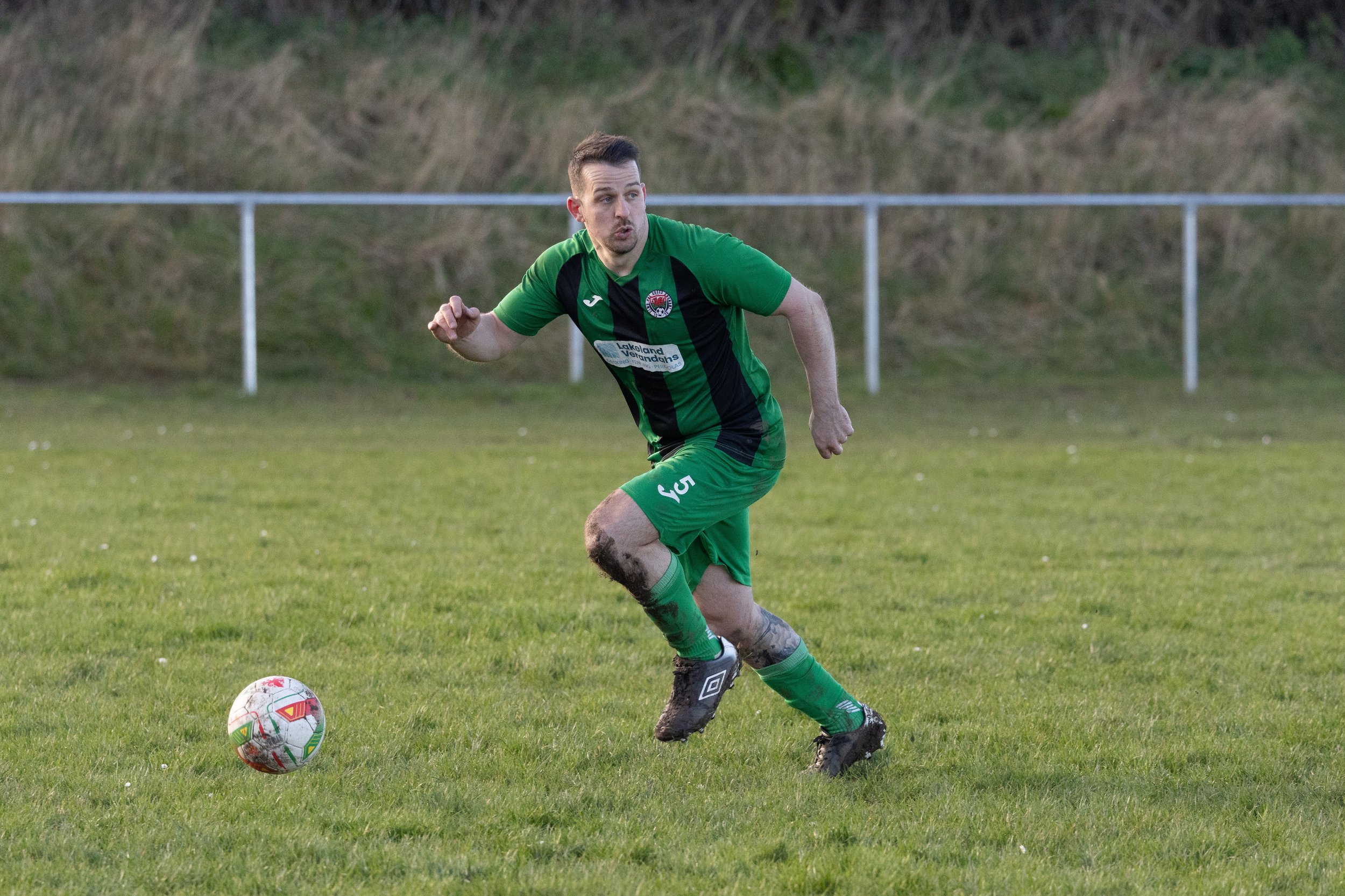A male soccer player wearing a green uniform with black accents kicking a soccer ball on a grassy field. His uniform has the number 5 and sports mud on his legs and shoes.