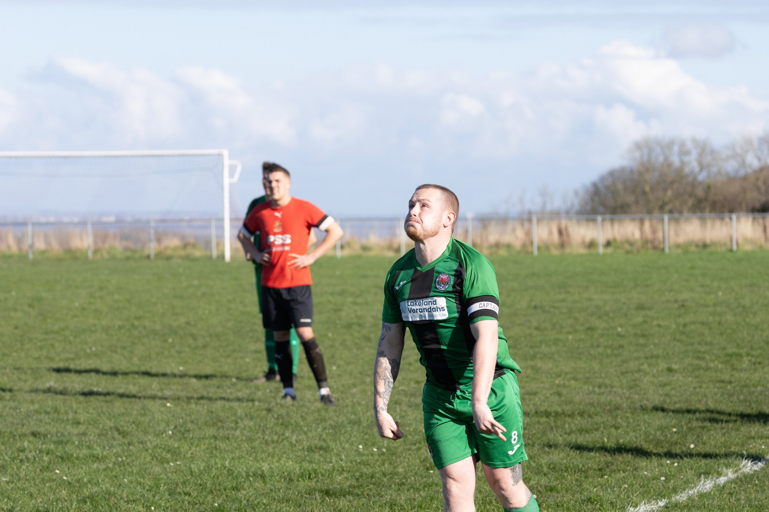 A soccer player in a green uniform, with tattoos on his arms, standing on the field with a disappointed expression, while two other players in red uniforms stand behind him under a cloudy sky.