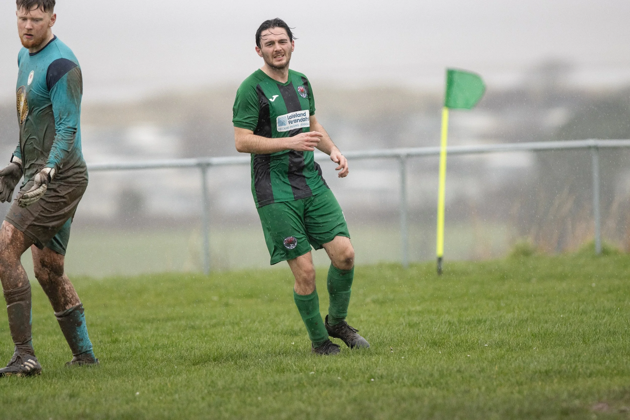 A soccer player in a green and black uniform stands on a rainy soccer field, near a corner flag, with another player in a muddy blue goalkeeper uniform nearby.