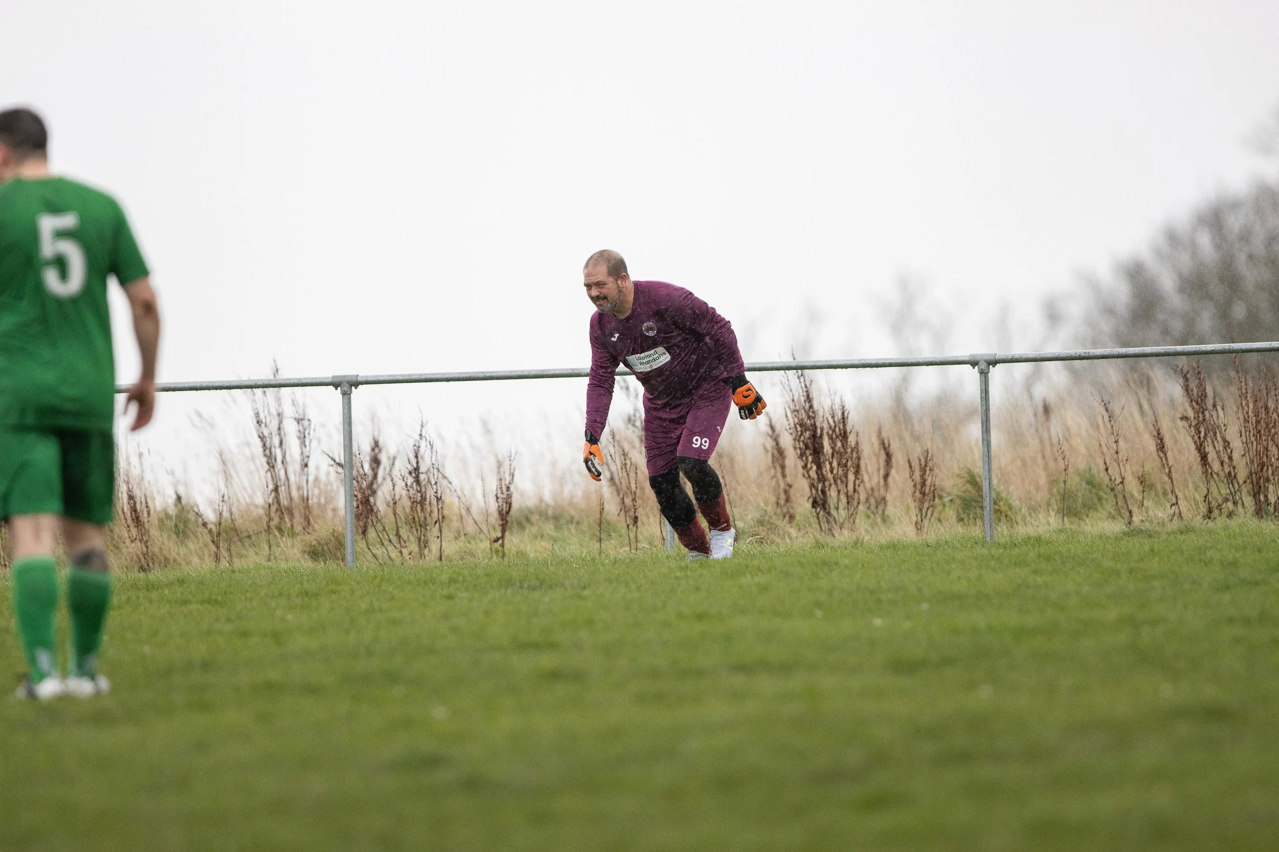 A soccer goalkeeper wearing a purple uniform and gloves is diving or falling on the field during a rainy game, with an out-of-focus opposing player in green and brown tall grass in the background.