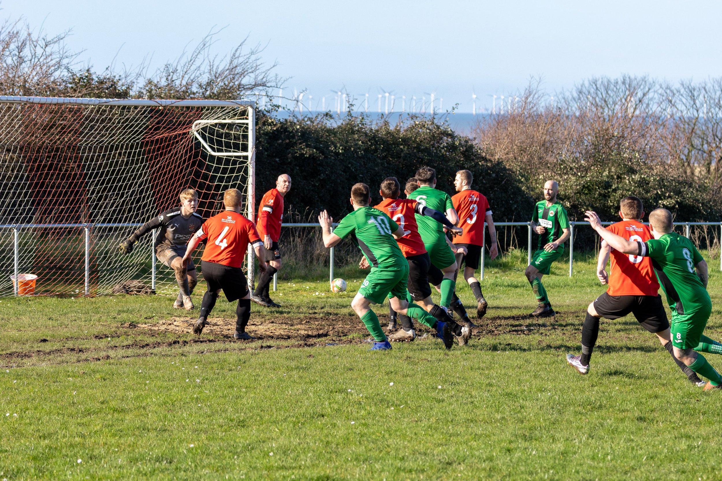 Soccer players in green and red jerseys compete for the ball near the goal on a grassy field, with a goalkeeper in black and officials in red nearby.