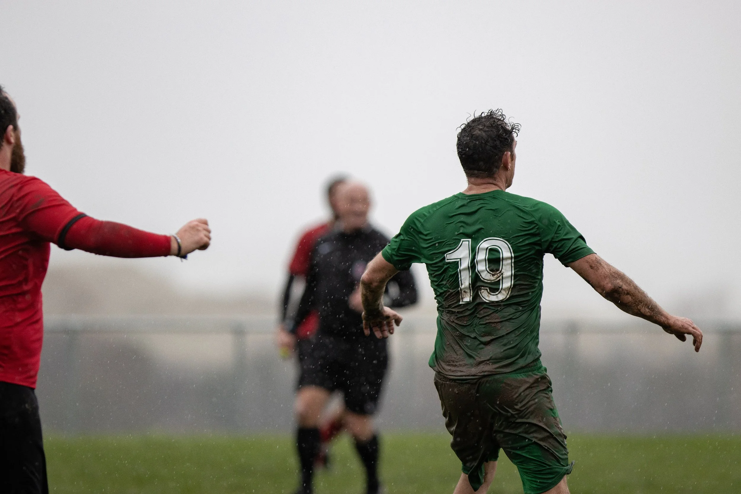 A soccer player wearing a green jersey with the number 19, covered in mud, runs on a wet field with two opponents in red jerseys and a referee in black and red in the background, on a cloudy, foggy day.