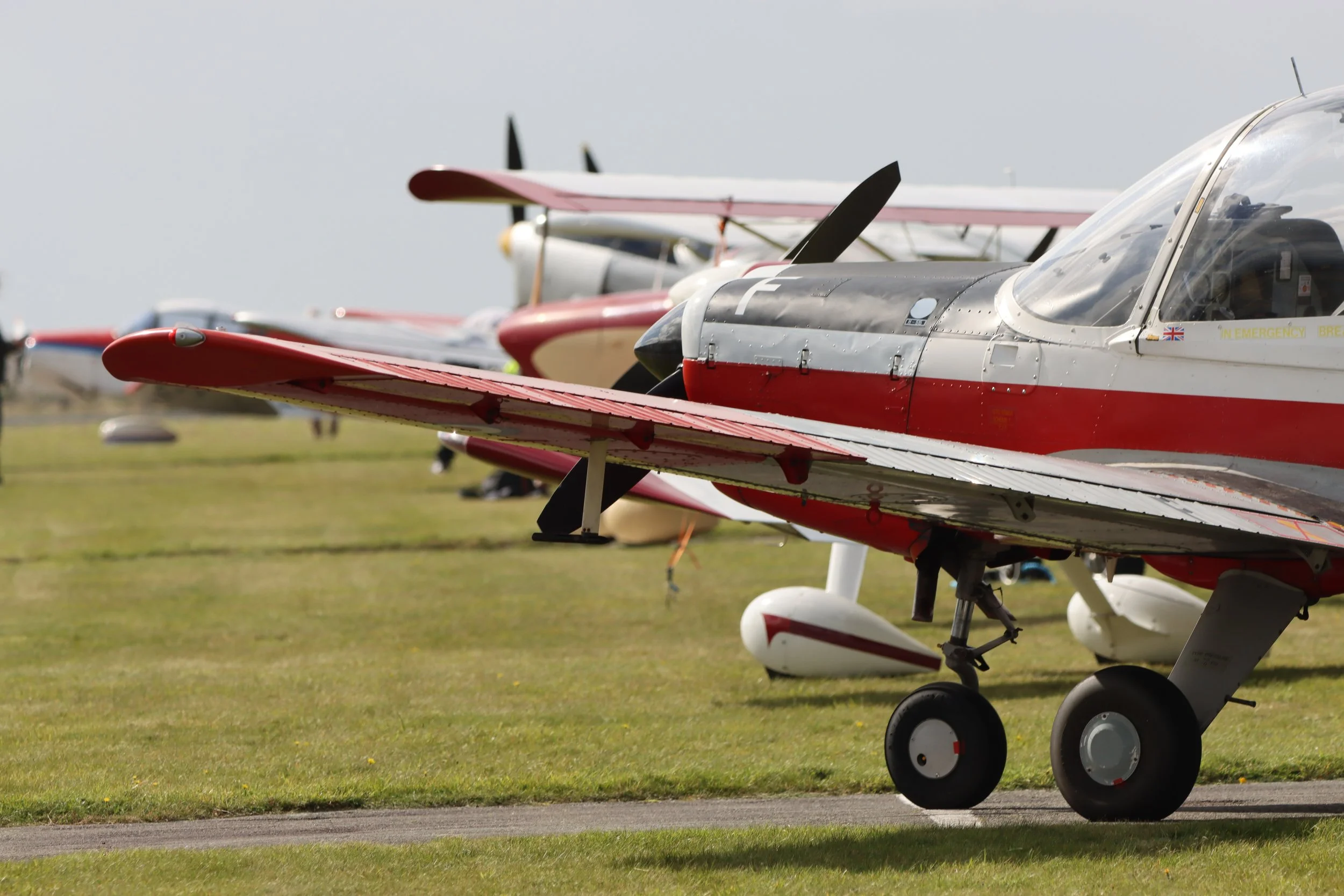 Multiple small aerobatic aircraft parked on a grassy airfield, with the focus on a red and white plane in the foreground.