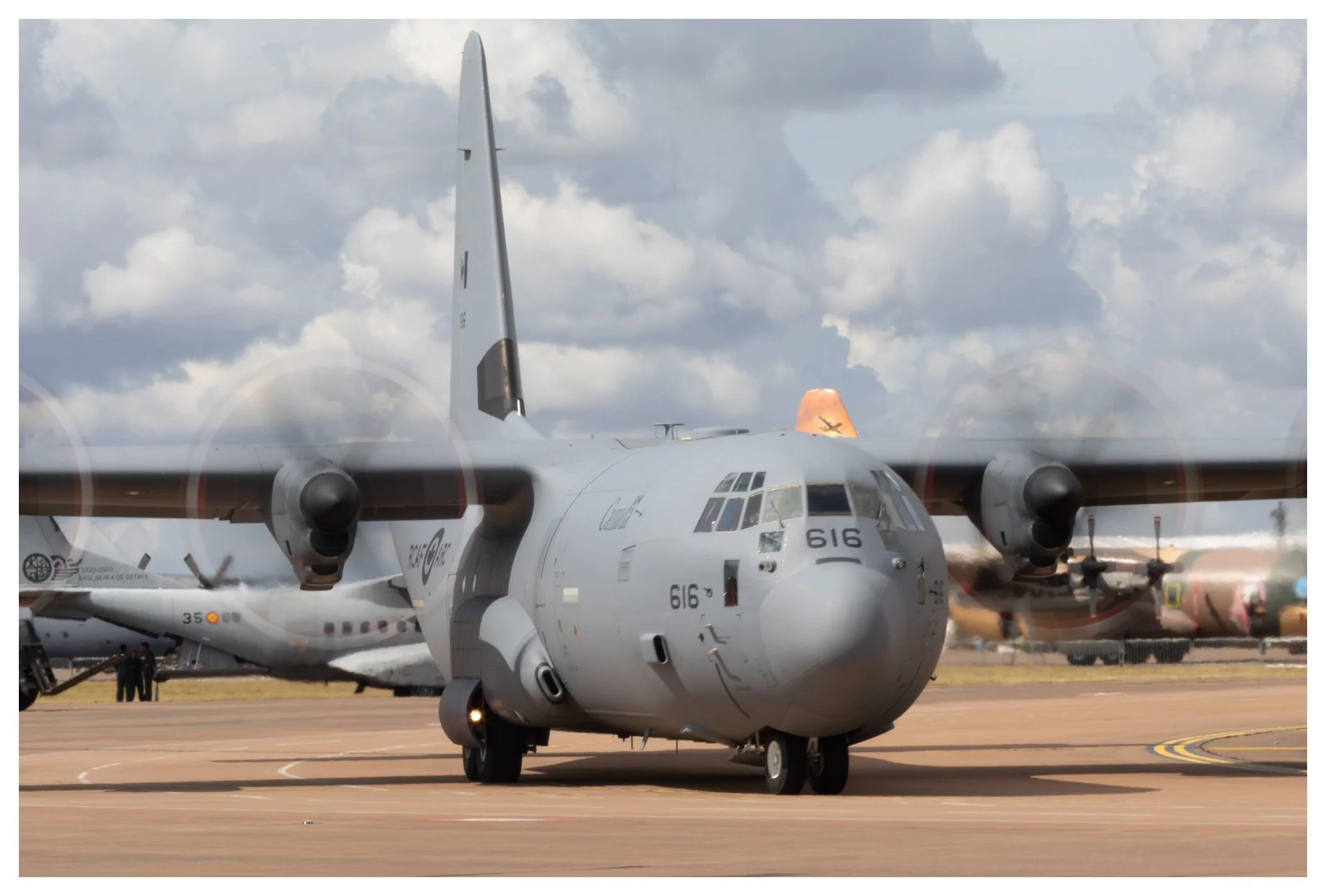 Military aircraft parked on an airfield, with other planes in the background and a partly cloudy sky.