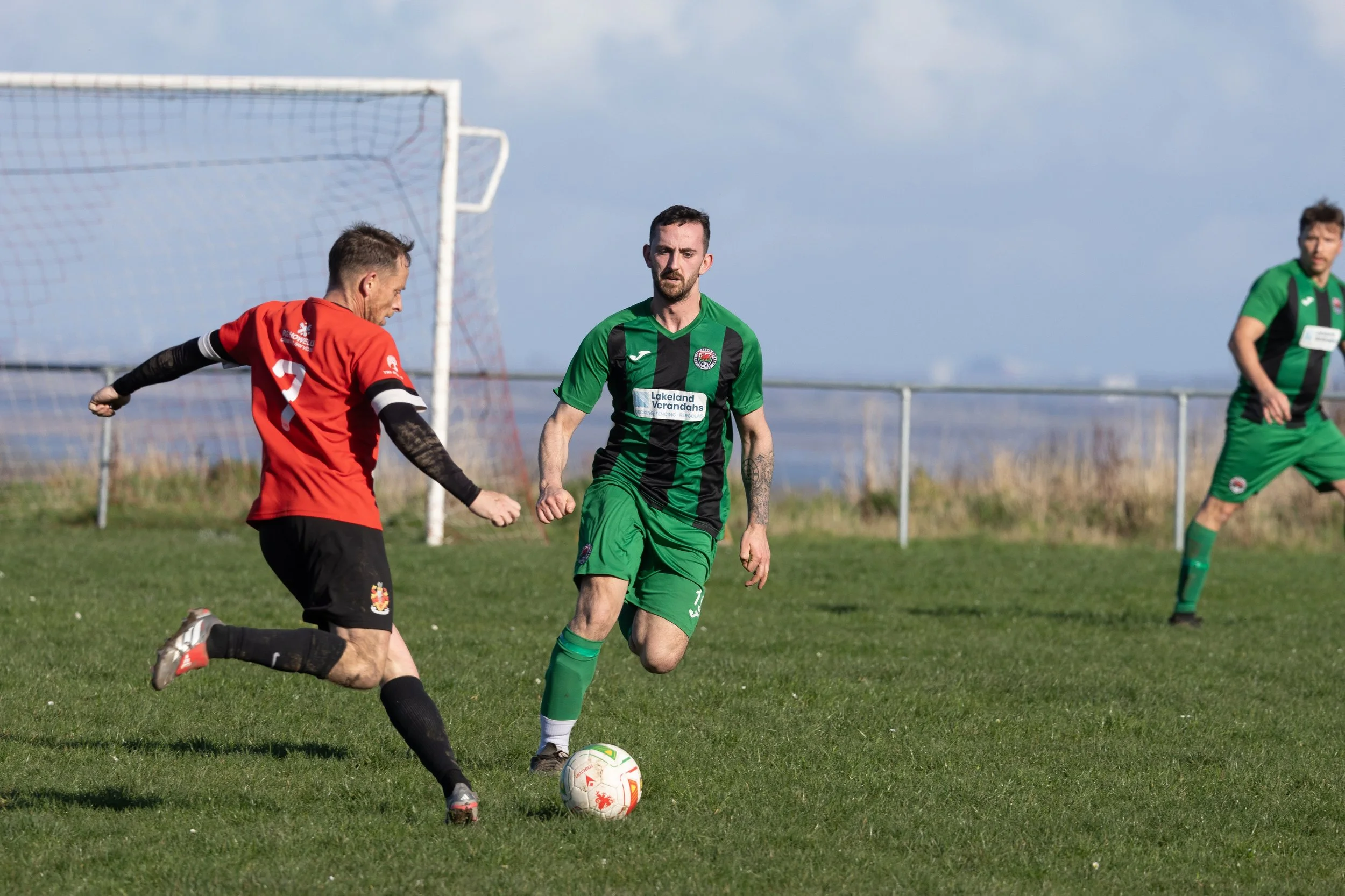 Soccer players in green and red uniforms competing for the ball on a grassy field, with goal net in background.