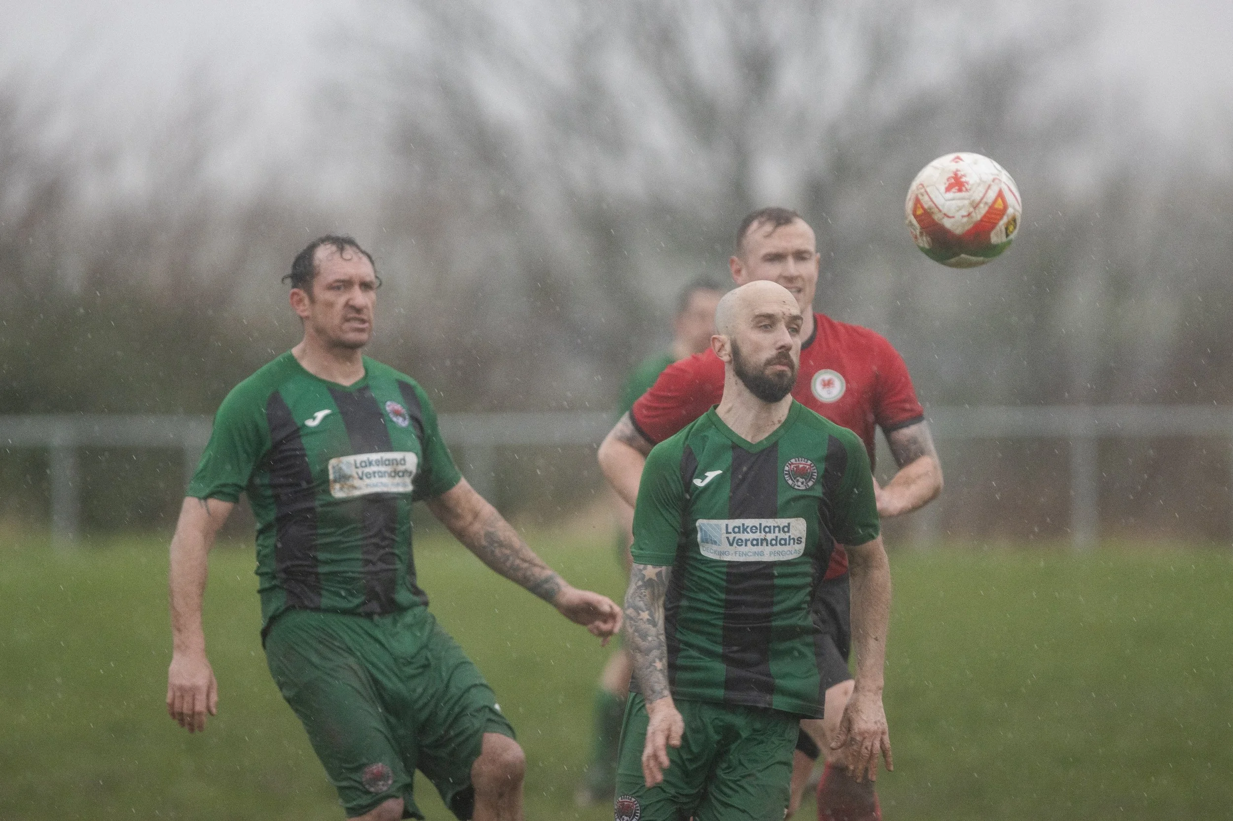 Soccer players in rain, wearing green and red uniforms, chasing a ball on a grassy field.