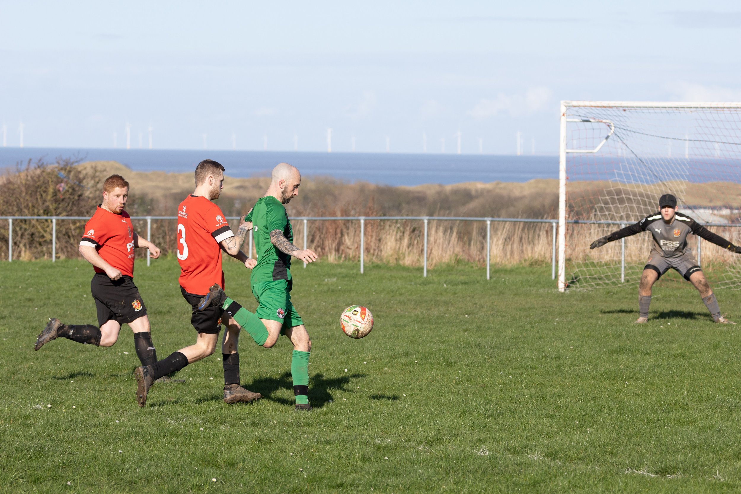 Three soccer players in red and green uniforms chasing the ball near the goal, with a goalkeeper in black preparing to block, against a background of wind turbines and a blue sky.