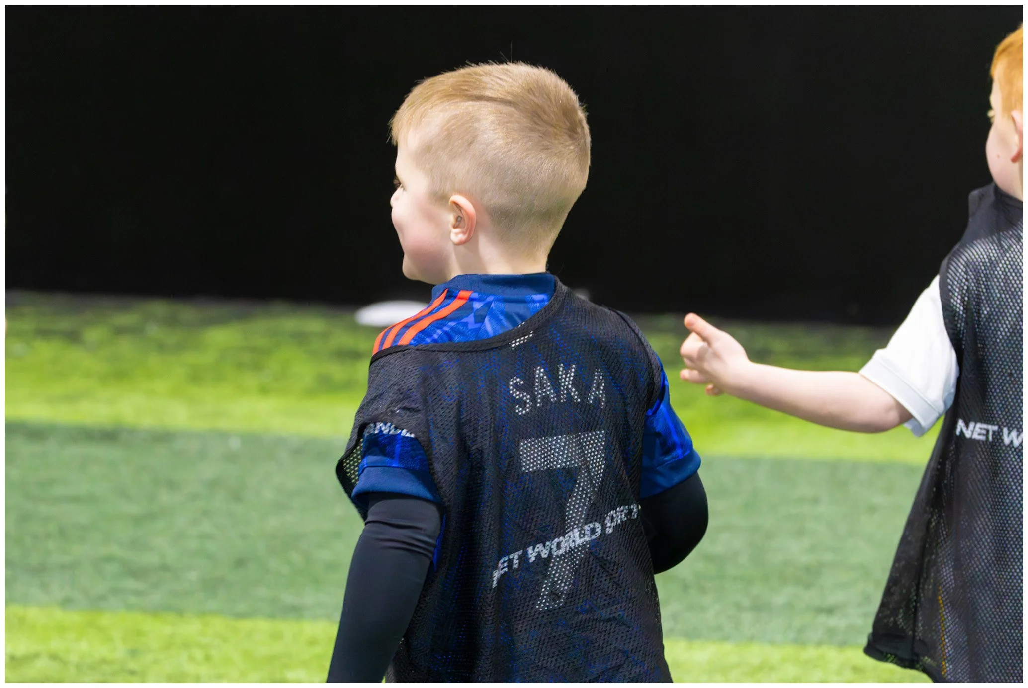 Two young boys wearing sports jerseys on a grass field, with one boy's jersey displaying the name 'Sakha' and the number 7 on the back.