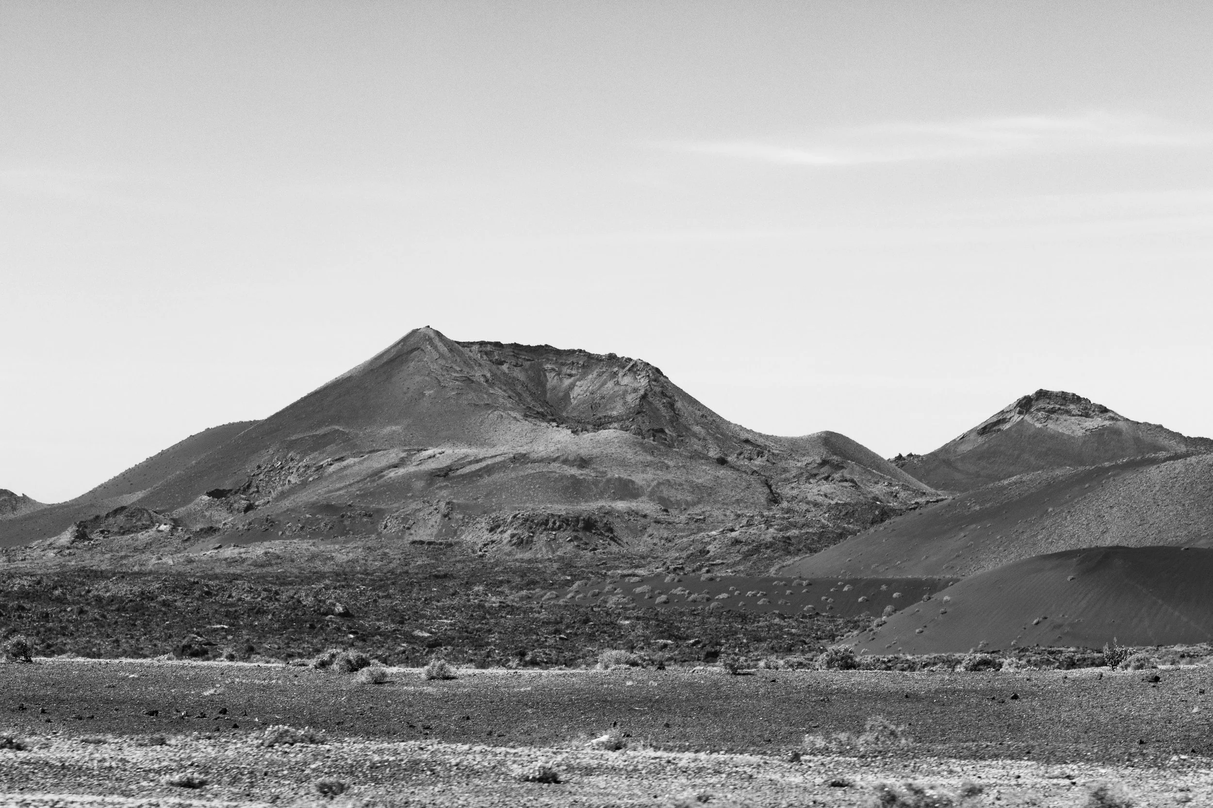 Black and white photo of a desert landscape with three mountains, sparse vegetation, and a clear sky.