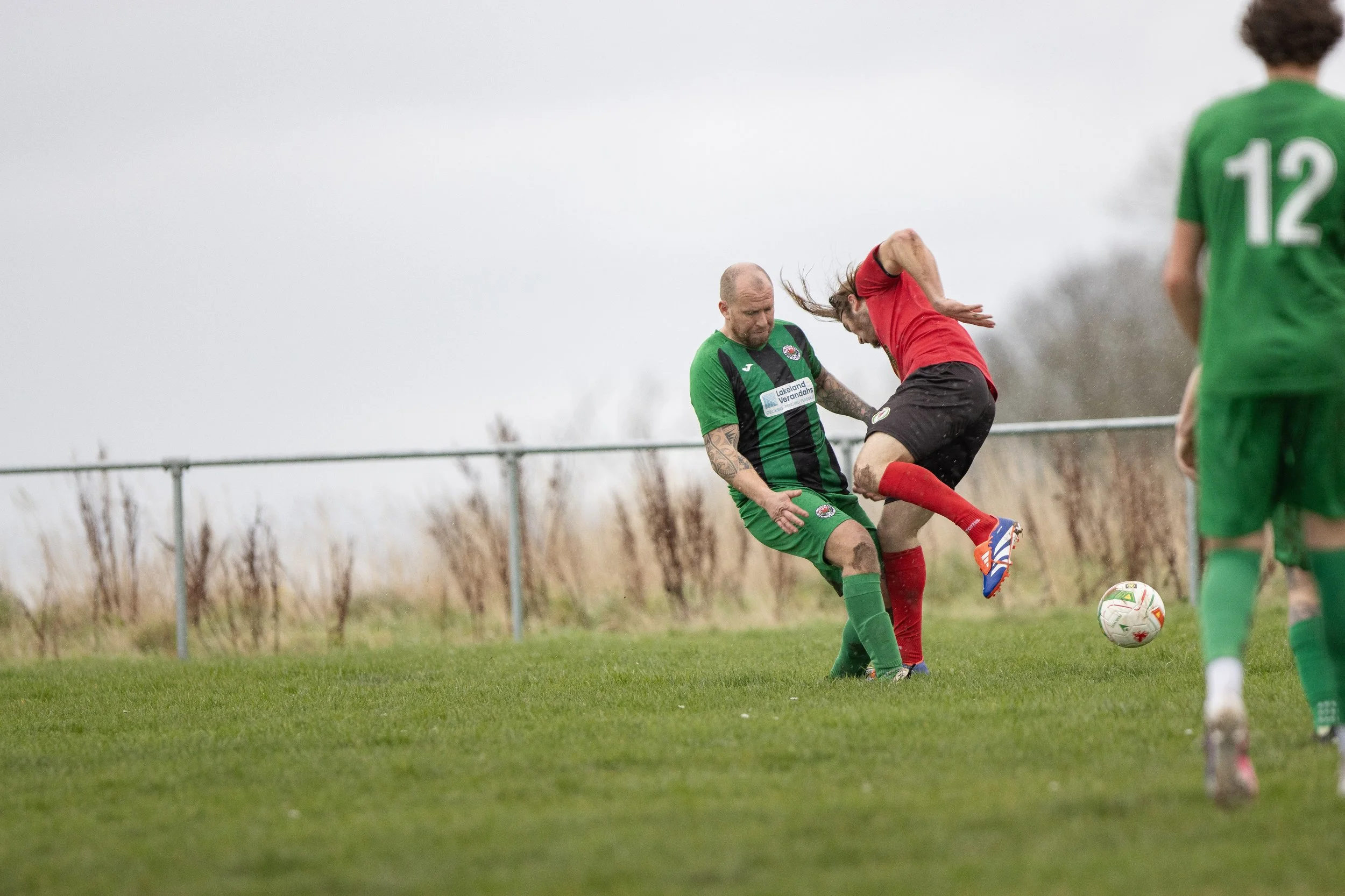 Two soccer players, one in a green uniform and the other in a red uniform, contest for the ball on a grassy field with a cloudy sky in the background. The player in red appears to be jumping or falling as the player in green blocks or challenges him.