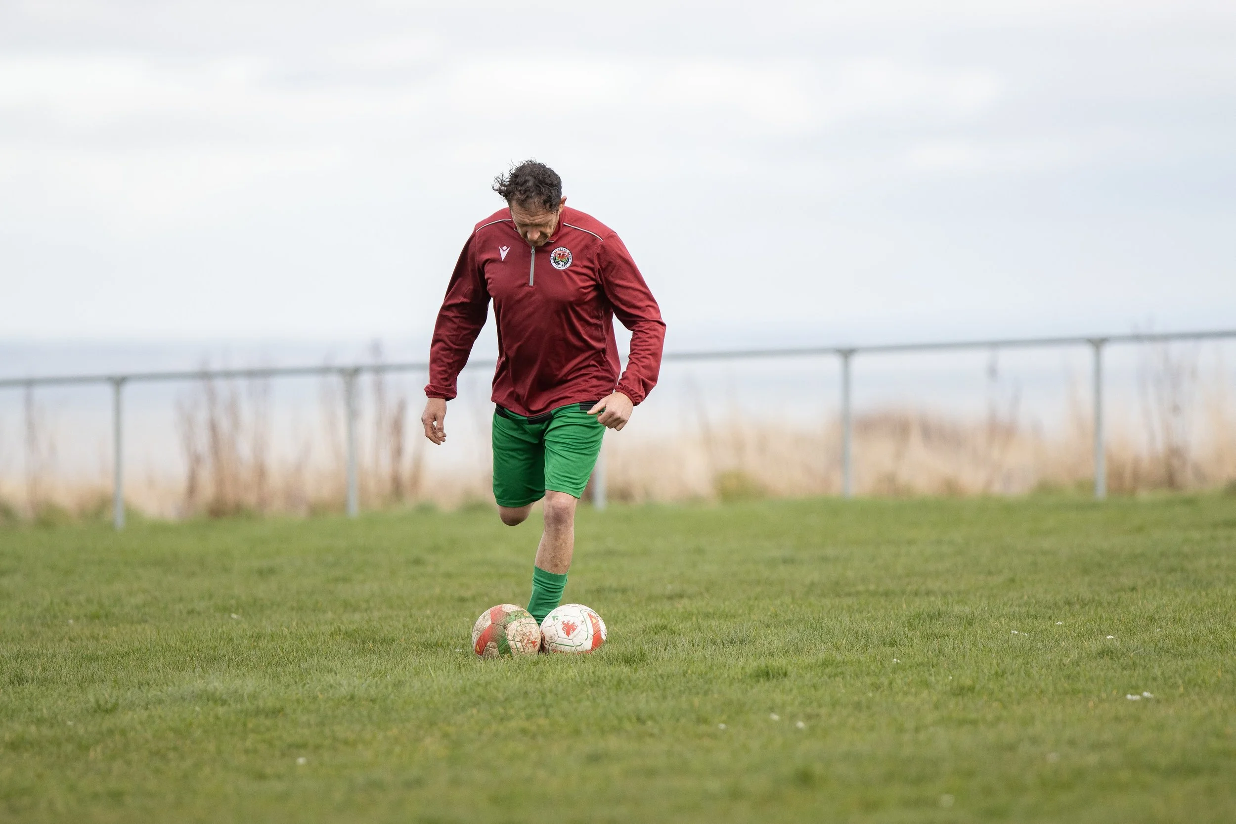 A man in a maroon jacket and green shorts kicks soccer balls on a grassy field.