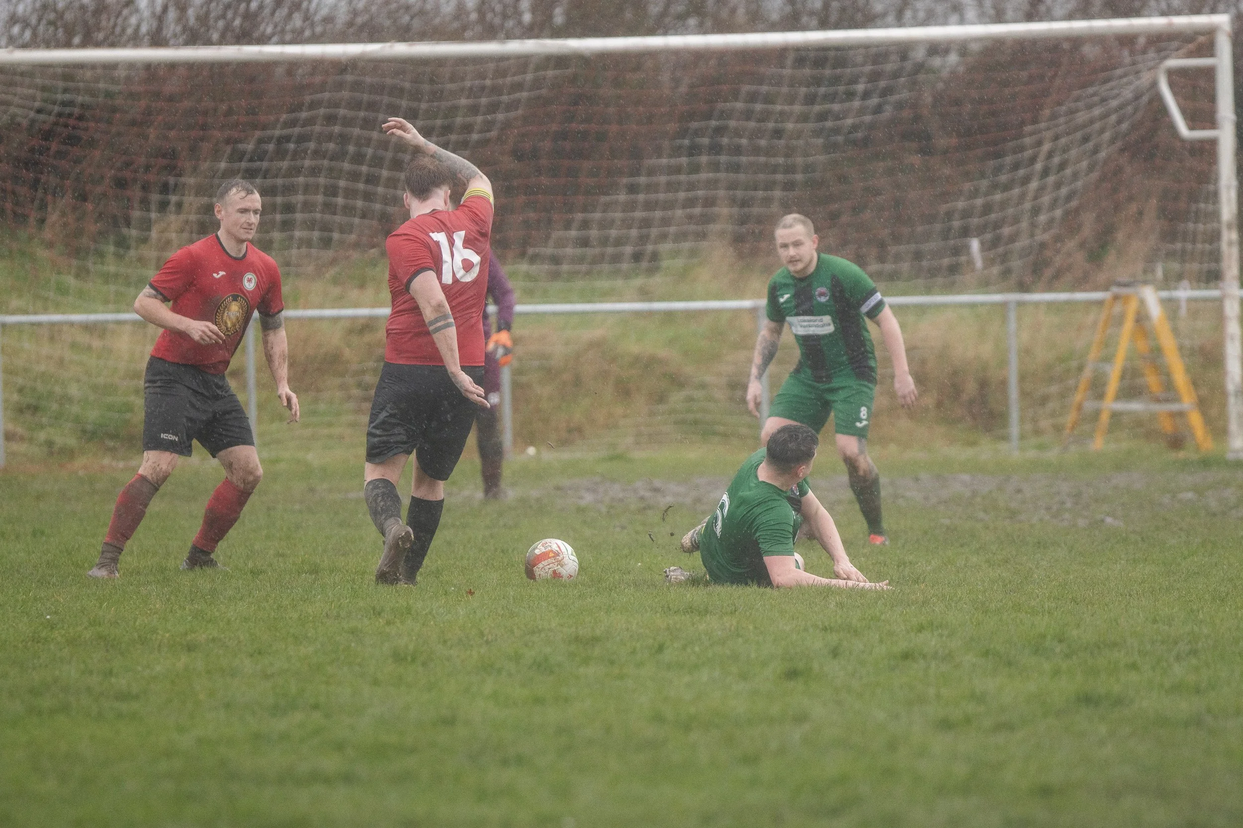 A group of soccer players on a muddy field during a game, with two players in red jerseys, two in green jerseys, and a rainy, foggy background.