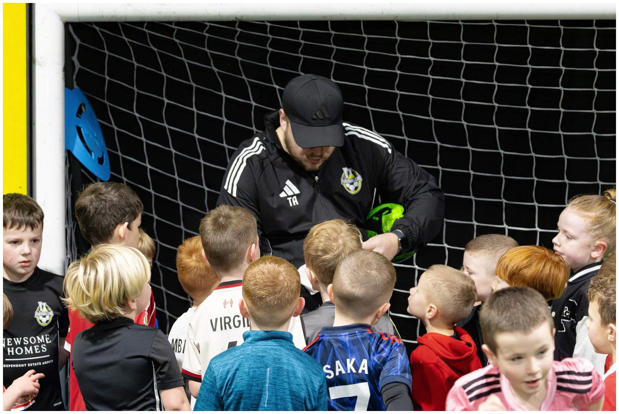 A group of young children gathered around a man in black sportswear, who is holding a green soccer helmet inside a goal net.