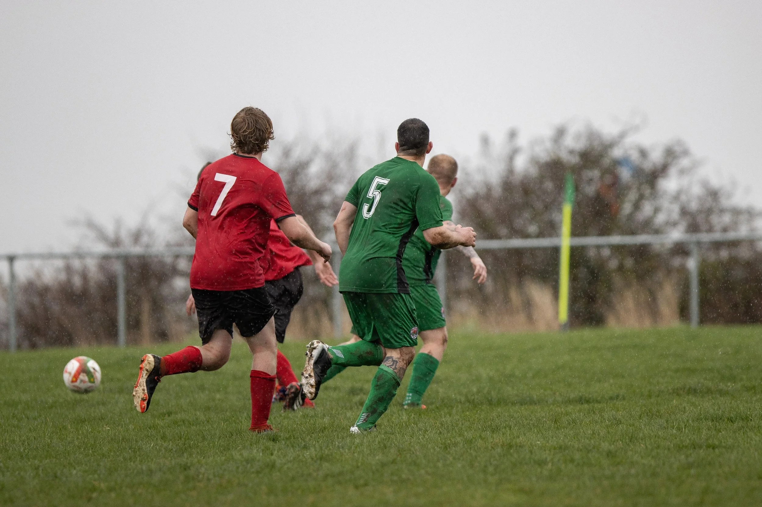 Soccer players in rain, wearing red and green uniforms, running on a grassy field near a corner flag, with wet grass and overcast sky.