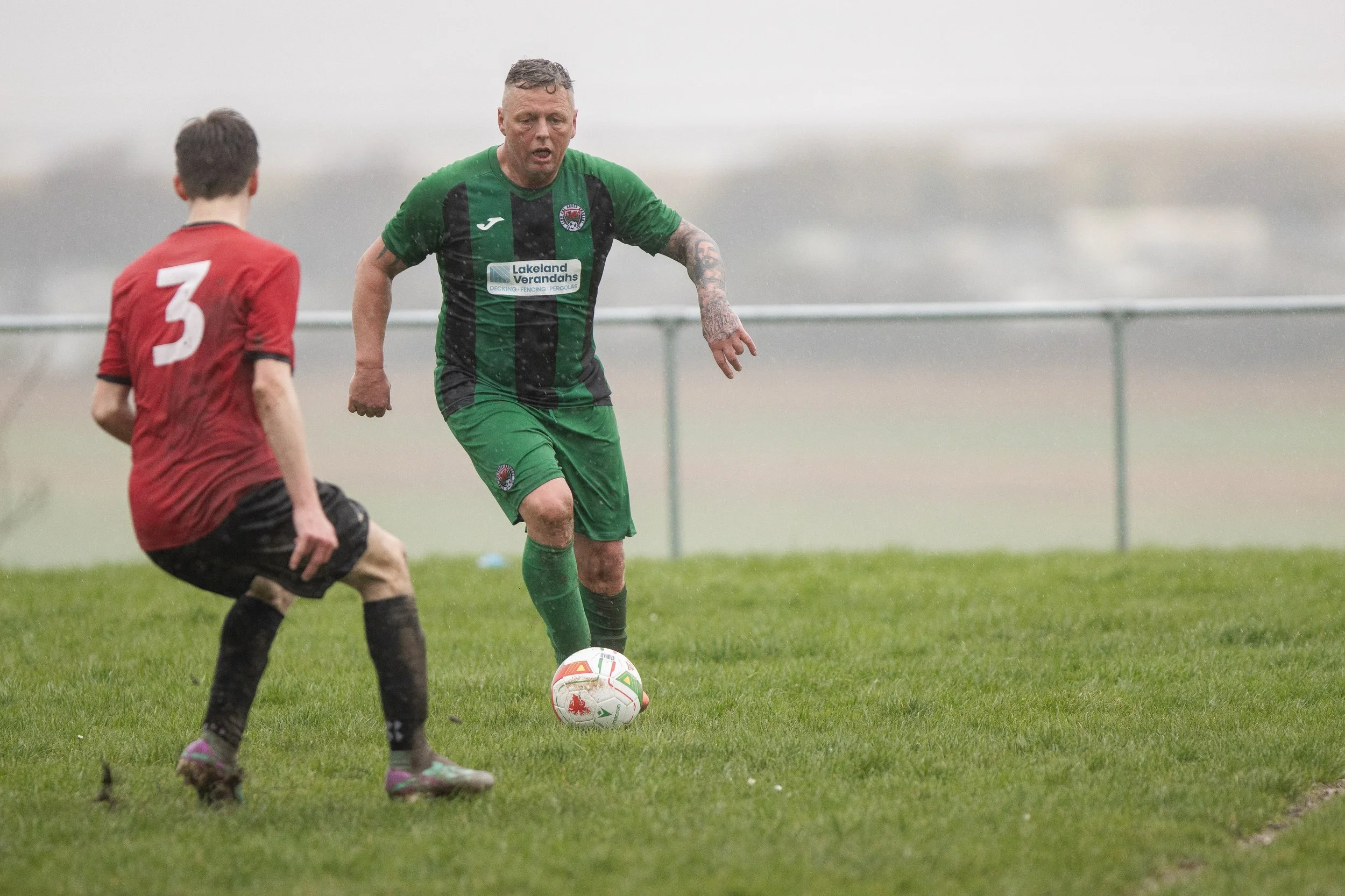 A soccer player in a green and black uniform preparing to kick the ball, while another player in a red and black uniform is nearby on a rainy day.