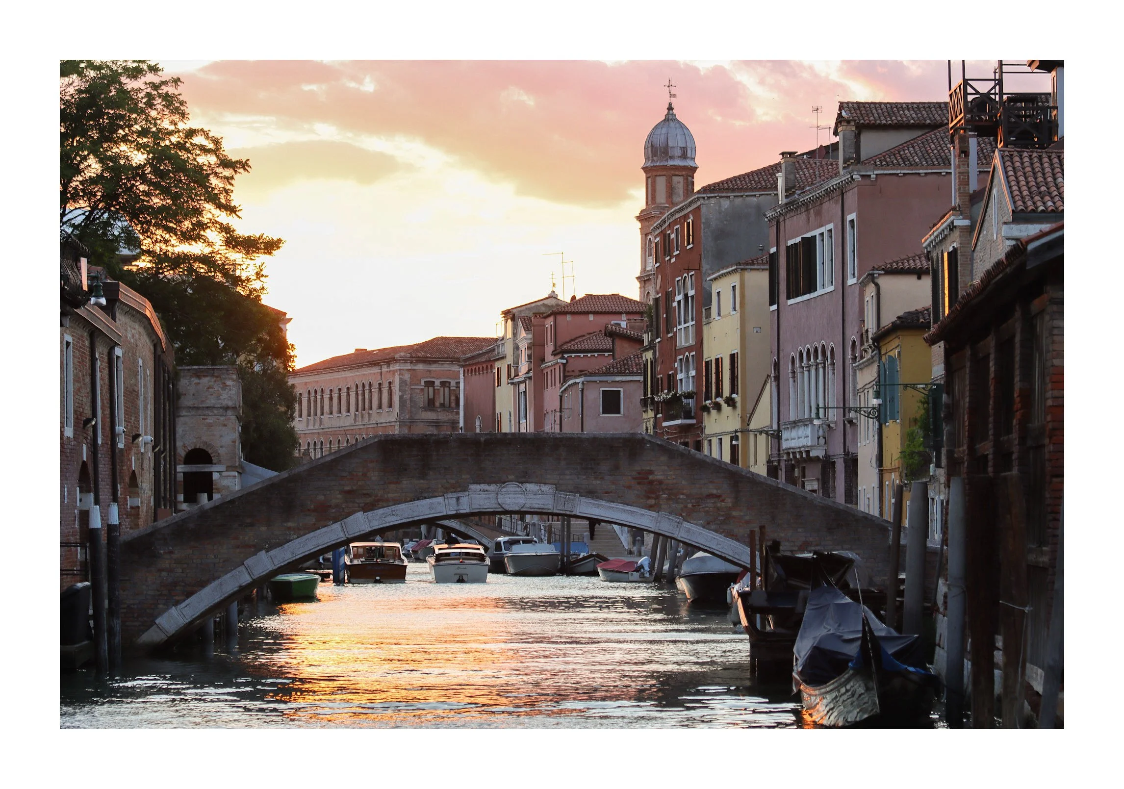 Sunset over a canal in Venice, Italy, with boats docked along the water and colorful old buildings lining the canal.