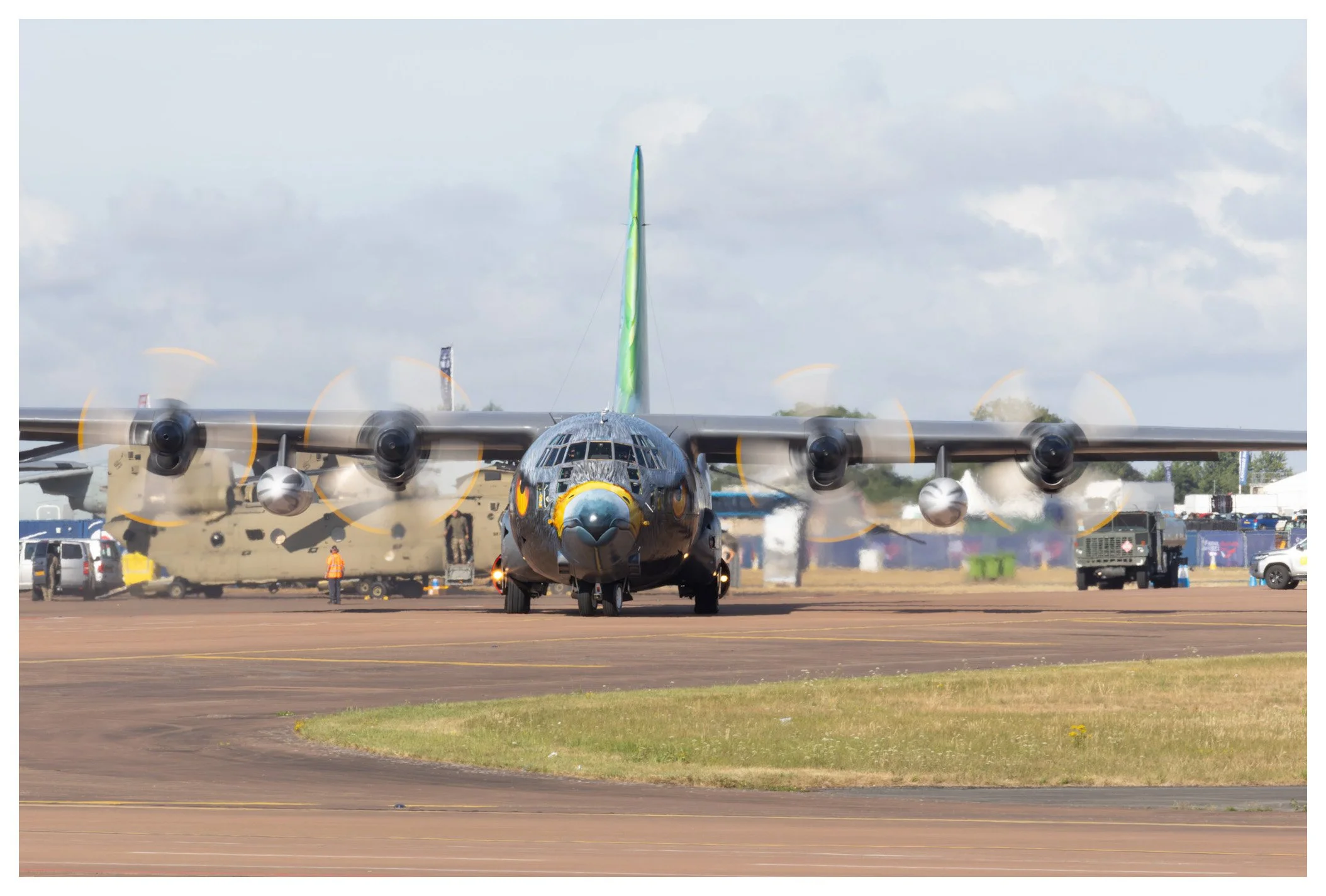 A military cargo airplane with four propeller engines on a runway, blurred motion from spinning propellers, with vehicles and personnel in the background, under a partly cloudy sky.