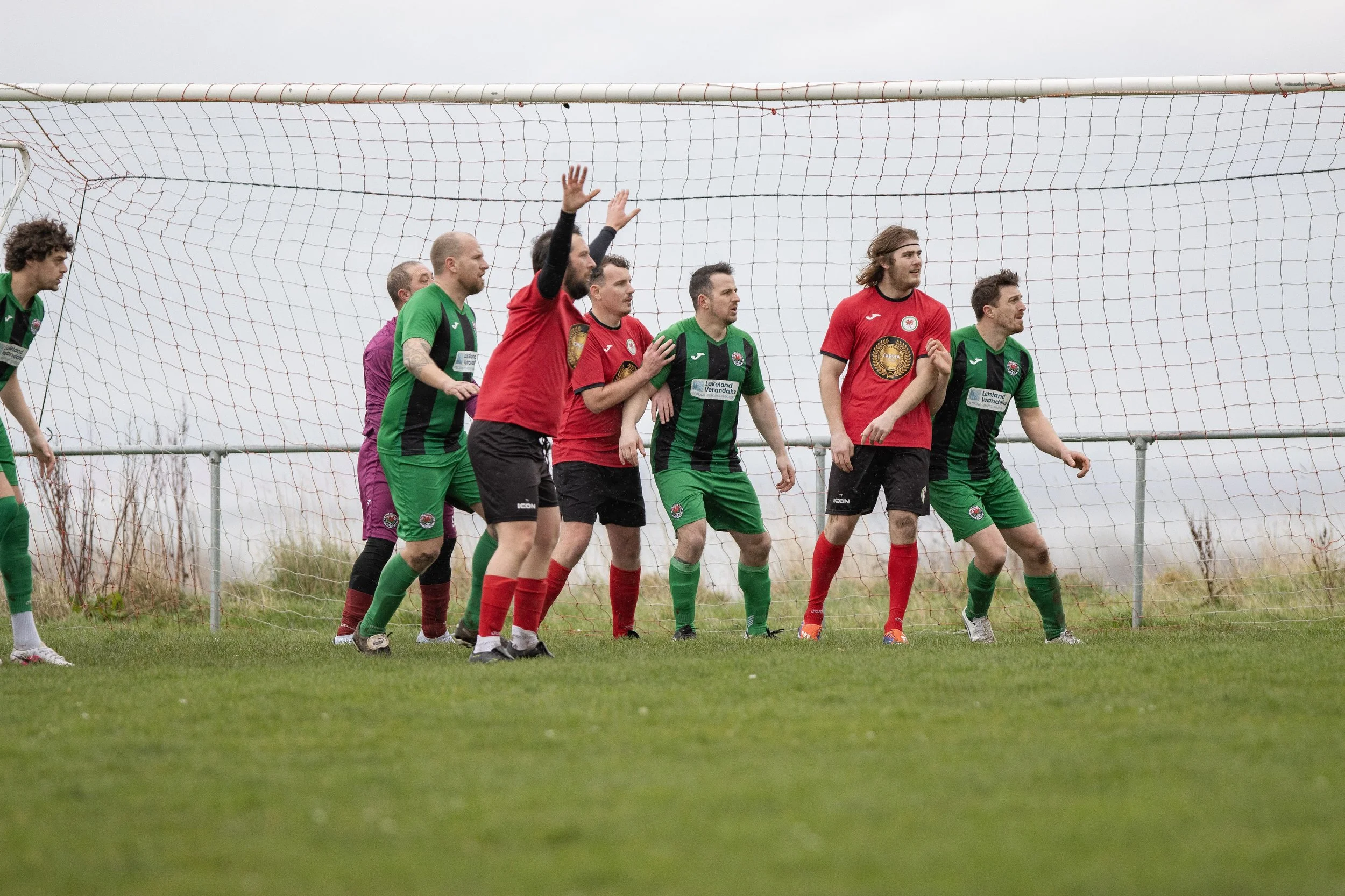 Soccer players in green and red jerseys preparing for a corner kick, standing near the goal in a grassy field.