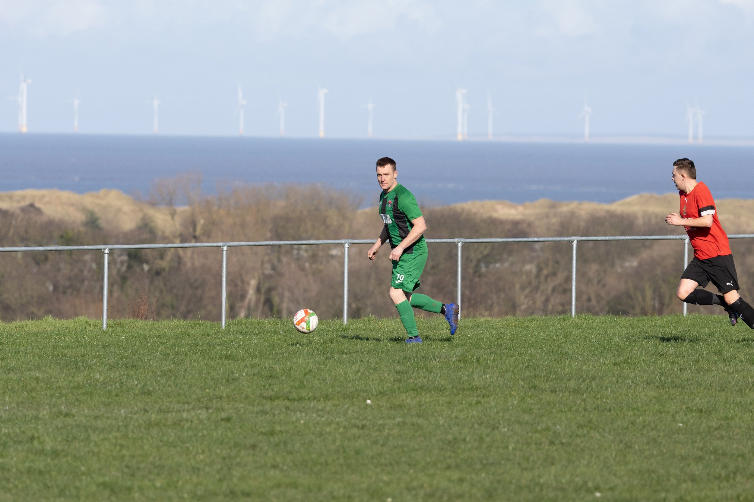Two soccer players, one in a green uniform and the other in a red uniform, running on a grass field with a metal fence and a scenic background of water and wind turbines.